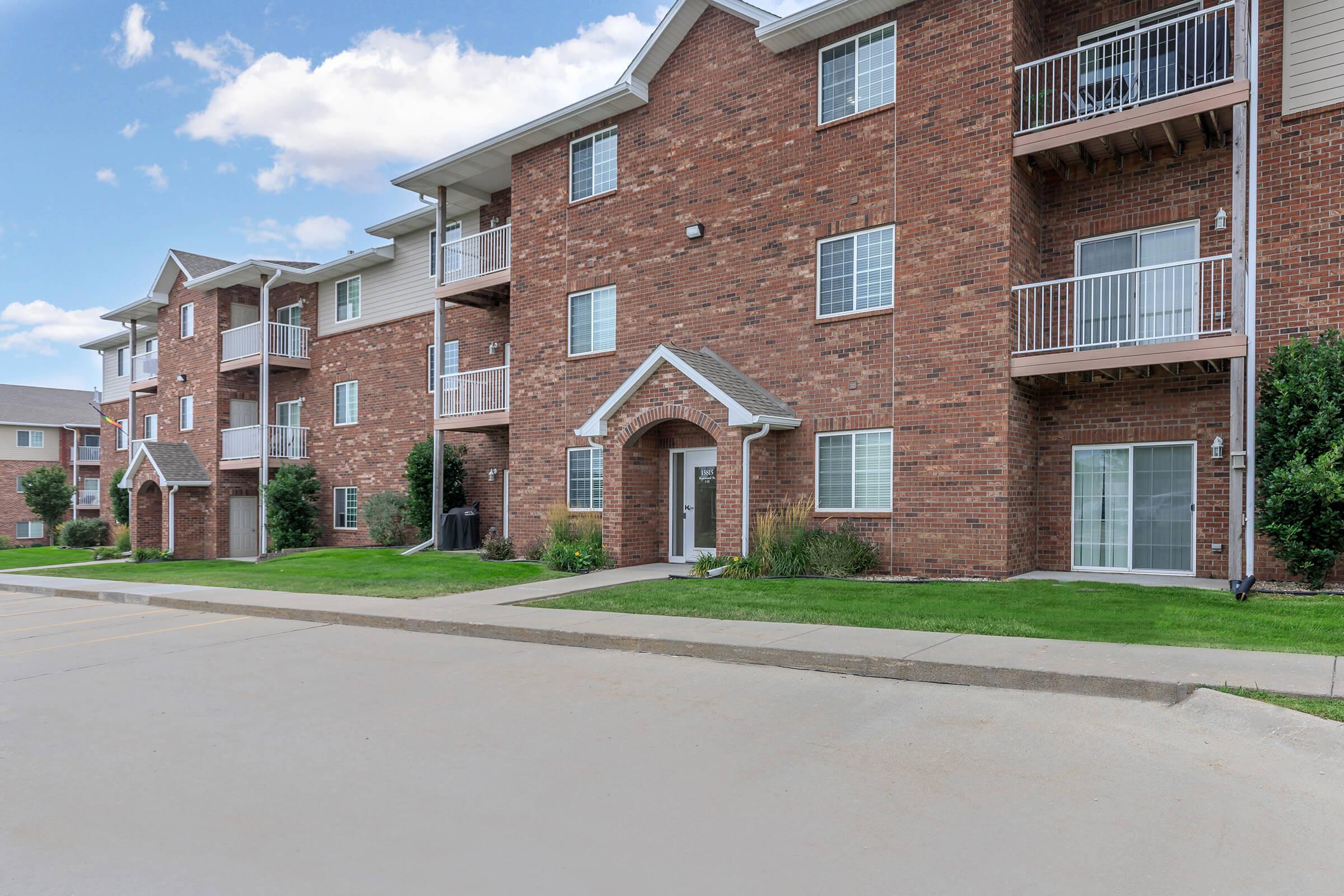 A brick apartment building with multiple units, featuring balconies and large windows. A well-maintained walkway leads to the main entrance, surrounded by green grass and small shrubs. The parking area is visible in the foreground, with a clear blue sky above.