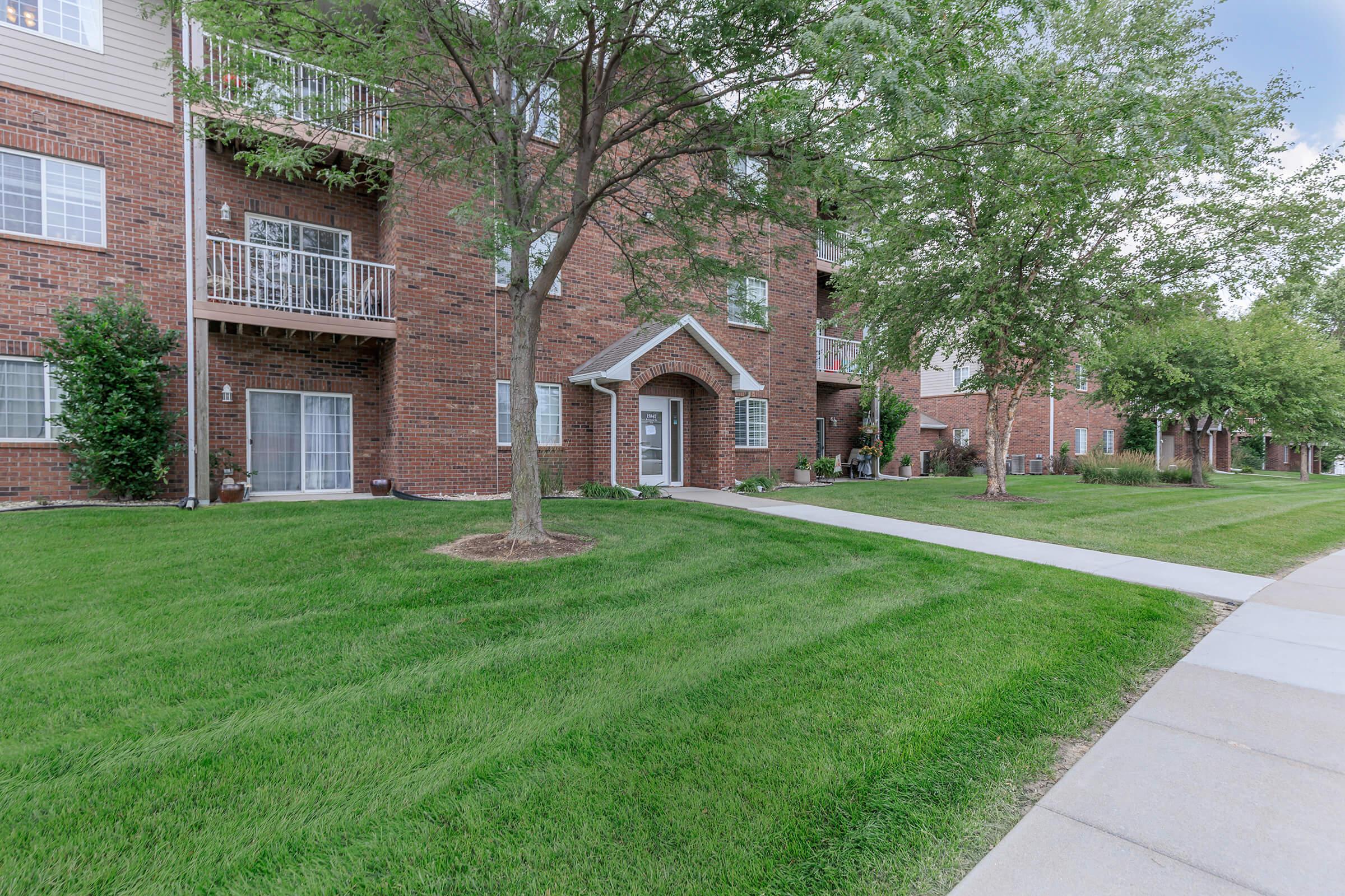 A well-maintained apartment building with red brick exterior. It features balconies, landscaped green grass, and trees in the foreground. A wide concrete pathway runs alongside the building, leading to the entrance, surrounded by neatly trimmed lawn.