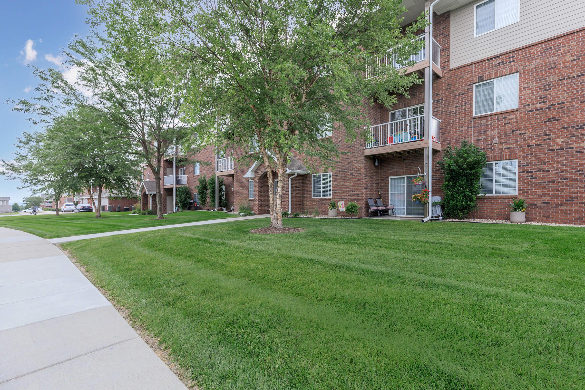 A well-maintained apartment complex with brick exteriors and green lawns. The image shows several units on the left, featuring balconies with plants and outdoor seating. Trees are planted along the walkway, which is paved, leading up to the buildings. The sky is clear with a few clouds.