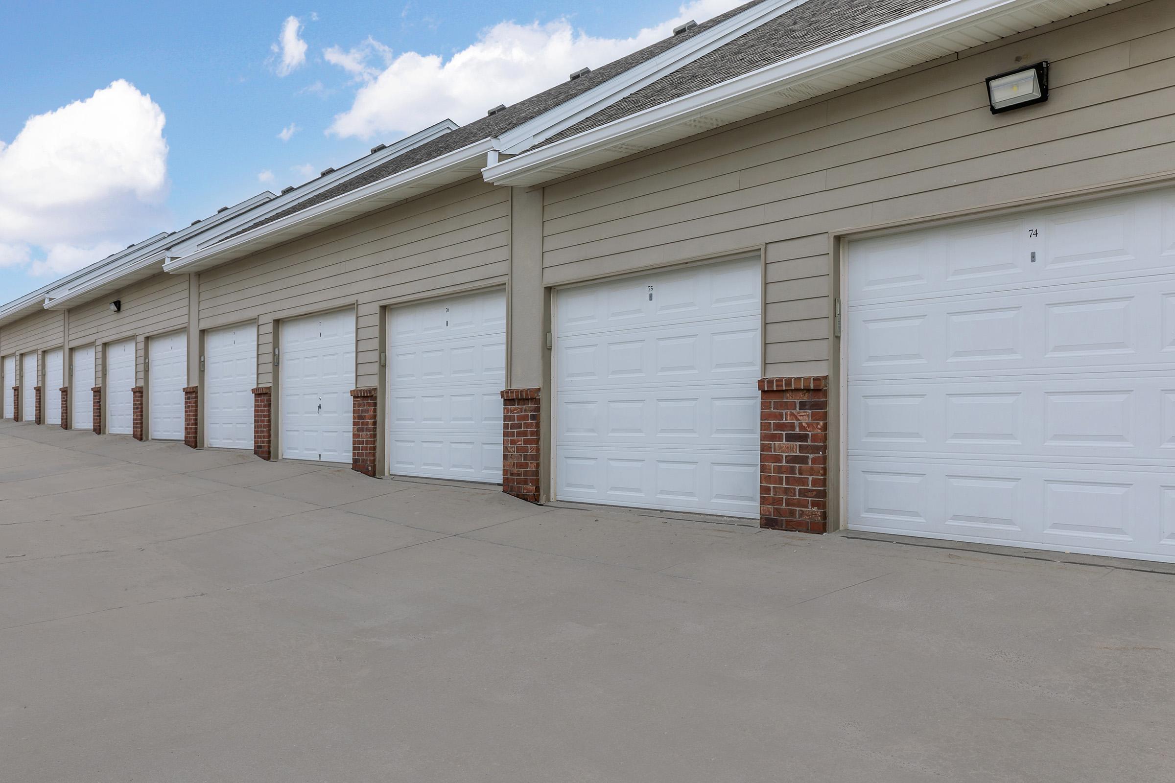 A row of garage doors painted white, with brick accents, set against a clear blue sky. The garages are arranged in a straight line, featuring numbers on each door. The driveway is paved and extends in front of the garages.
