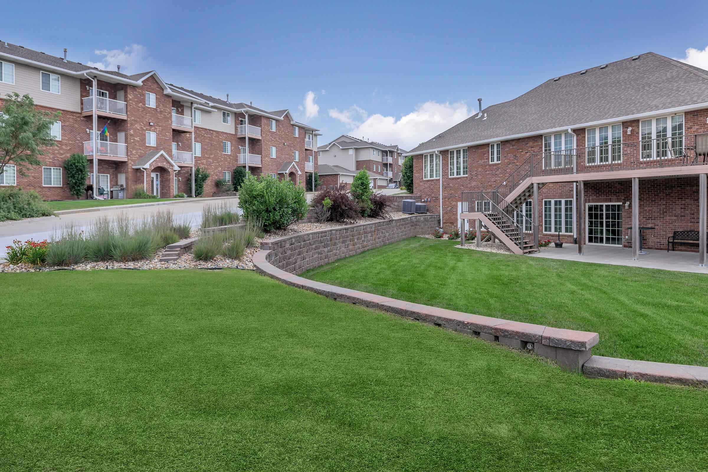 A well-maintained suburban area featuring a lush green lawn, landscaped garden beds, and two multi-story brick apartment buildings in the background. The scene is bright and inviting, showcasing outdoor seating on a patio and a paved walkway leading up to the buildings.