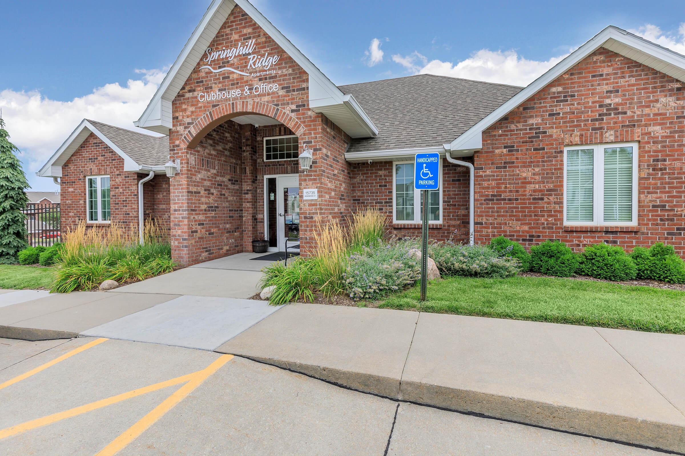 A brick building with a gabled roof labeled "Springhill Ridge Clubhouse & Office." The entrance features a covered porch with large windows. The surrounding area has well-maintained landscaping with grass, shrubs, and a sign indicating accessible parking. A clear blue sky is visible above.