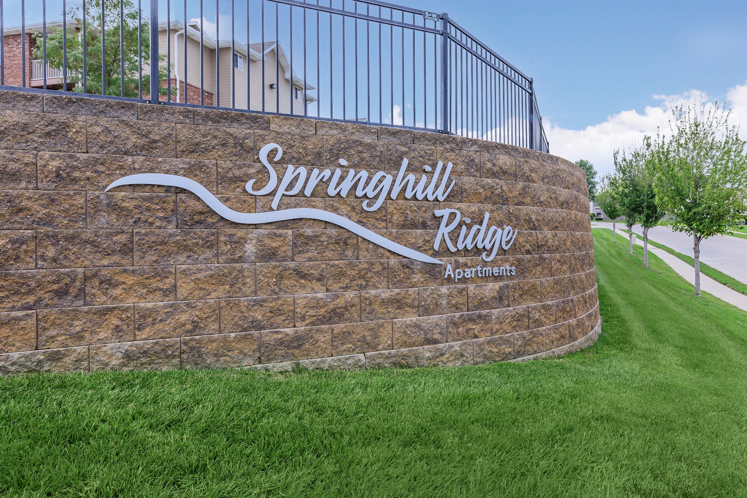 Signage for Springhill Ridge Apartments on a stone wall, surrounded by green grass and featuring a clear blue sky in the background. The sign includes the name "Springhill Ridge" in stylish lettering, along with the word "Apartments."