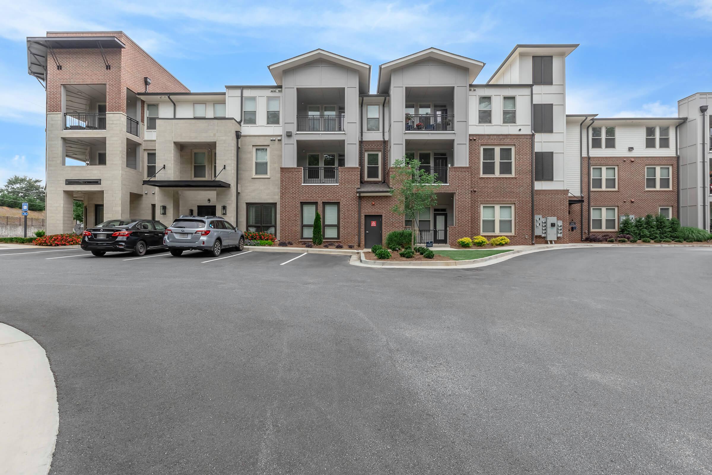 A modern apartment building featuring a mix of brick and siding exterior with multiple stories. The front area includes parking spaces for cars and landscaped flowerbeds. The design showcases contemporary architecture with large windows and balconies.