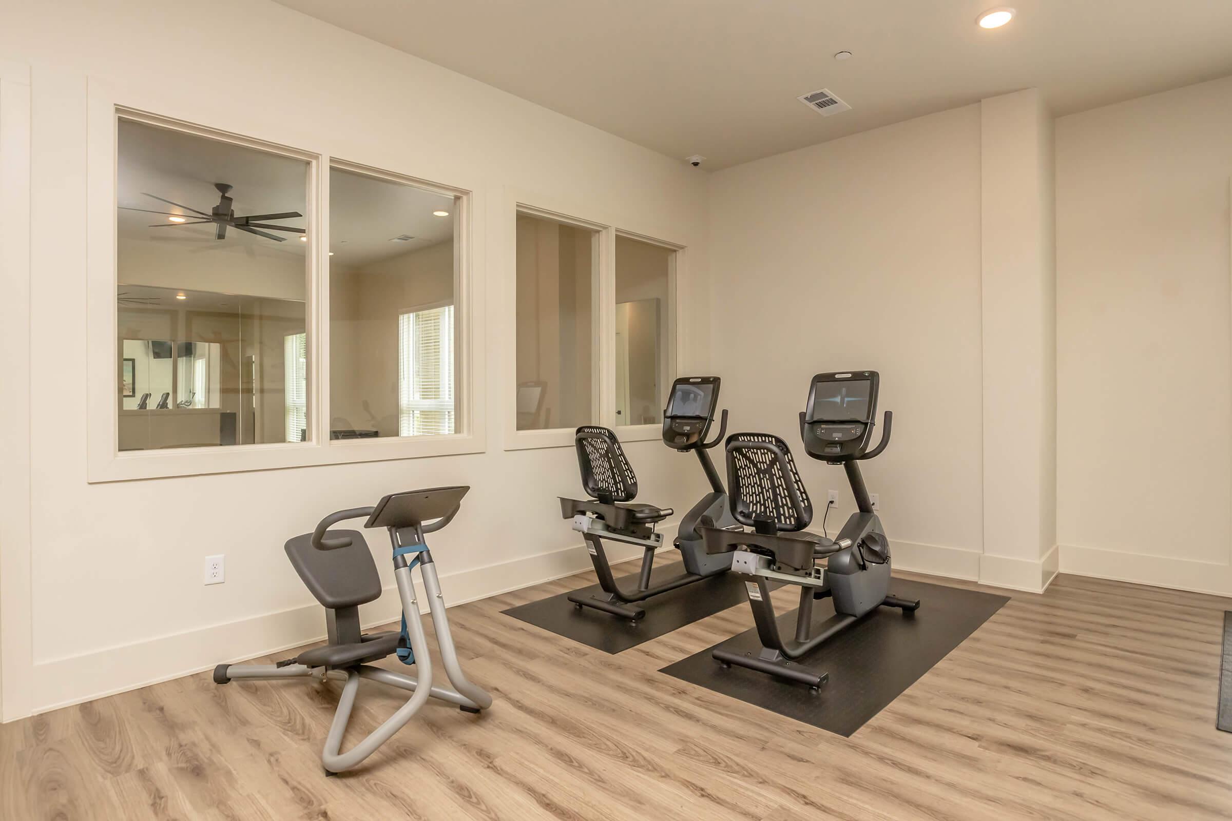 A bright, spacious fitness room with three exercise machines on a wooden floor. Large windows allow natural light to flood the space, and modern lighting fixtures enhance the ambiance. The walls are painted in light colors, creating an inviting atmosphere for workouts.