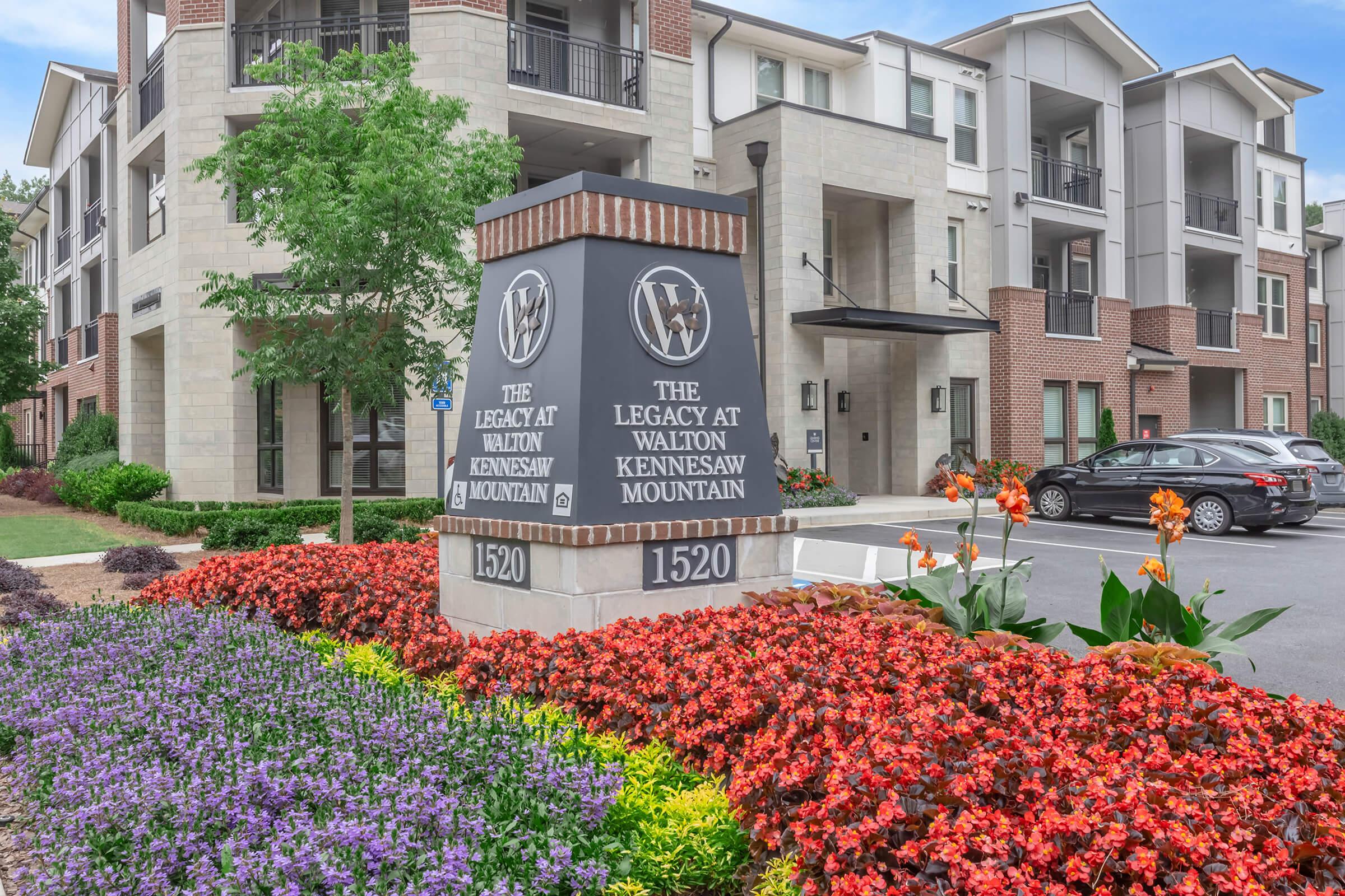 Sign at the entrance of an apartment complex, "The Legacy at Walton Kennesaw Mountain," displaying the address "1520." The sign is surrounded by vibrant flower beds filled with orange and purple flowers, and modern apartment buildings are visible in the background. Clear blue sky overhead.