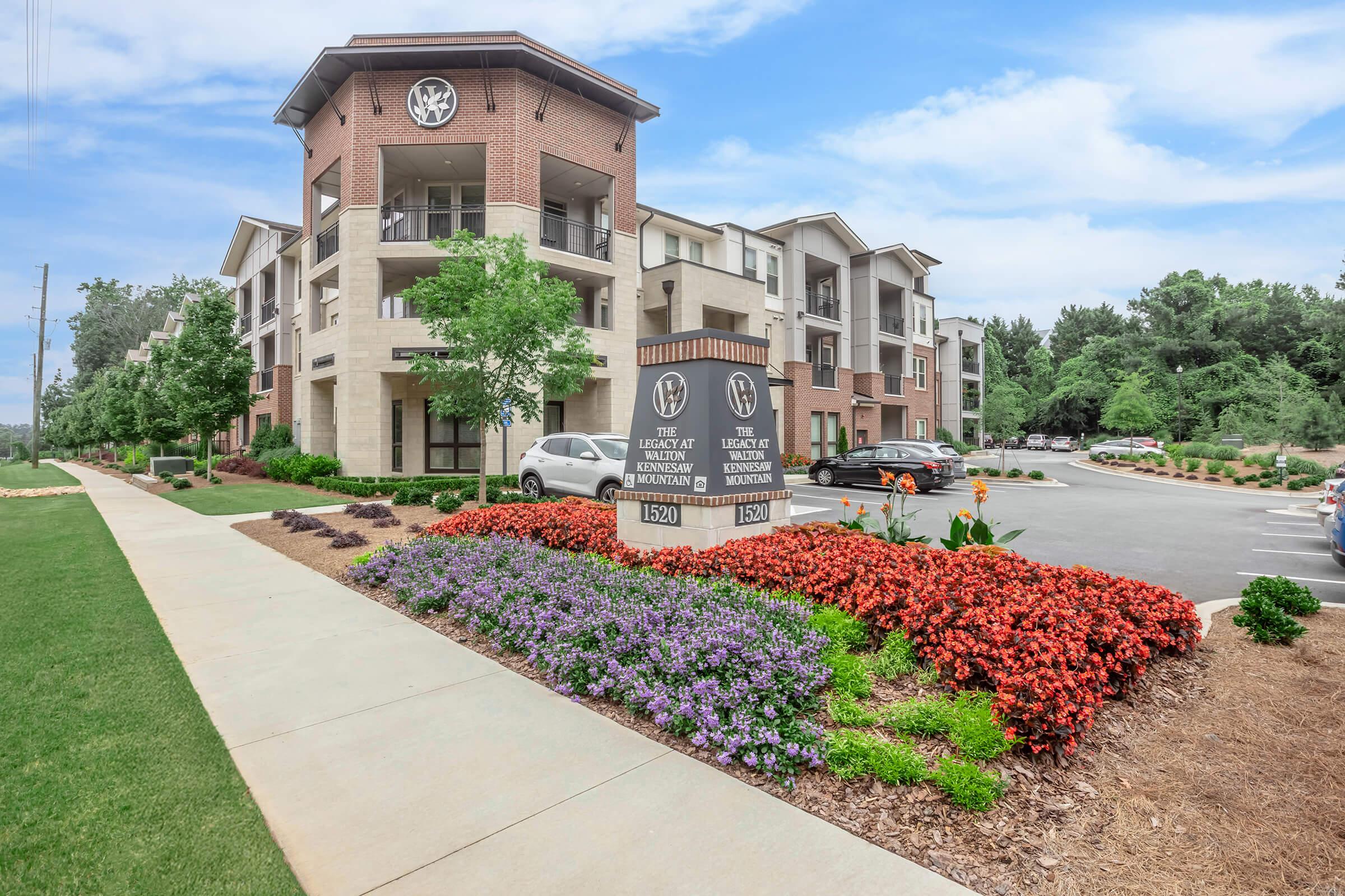 Modern brick apartment building with landscaped flowerbeds featuring colorful blossoms. A prominent sign indicates the property name and address, surrounded by well-maintained greenery. There are parked cars and a clear sky in the background, showcasing a welcoming residential environment.