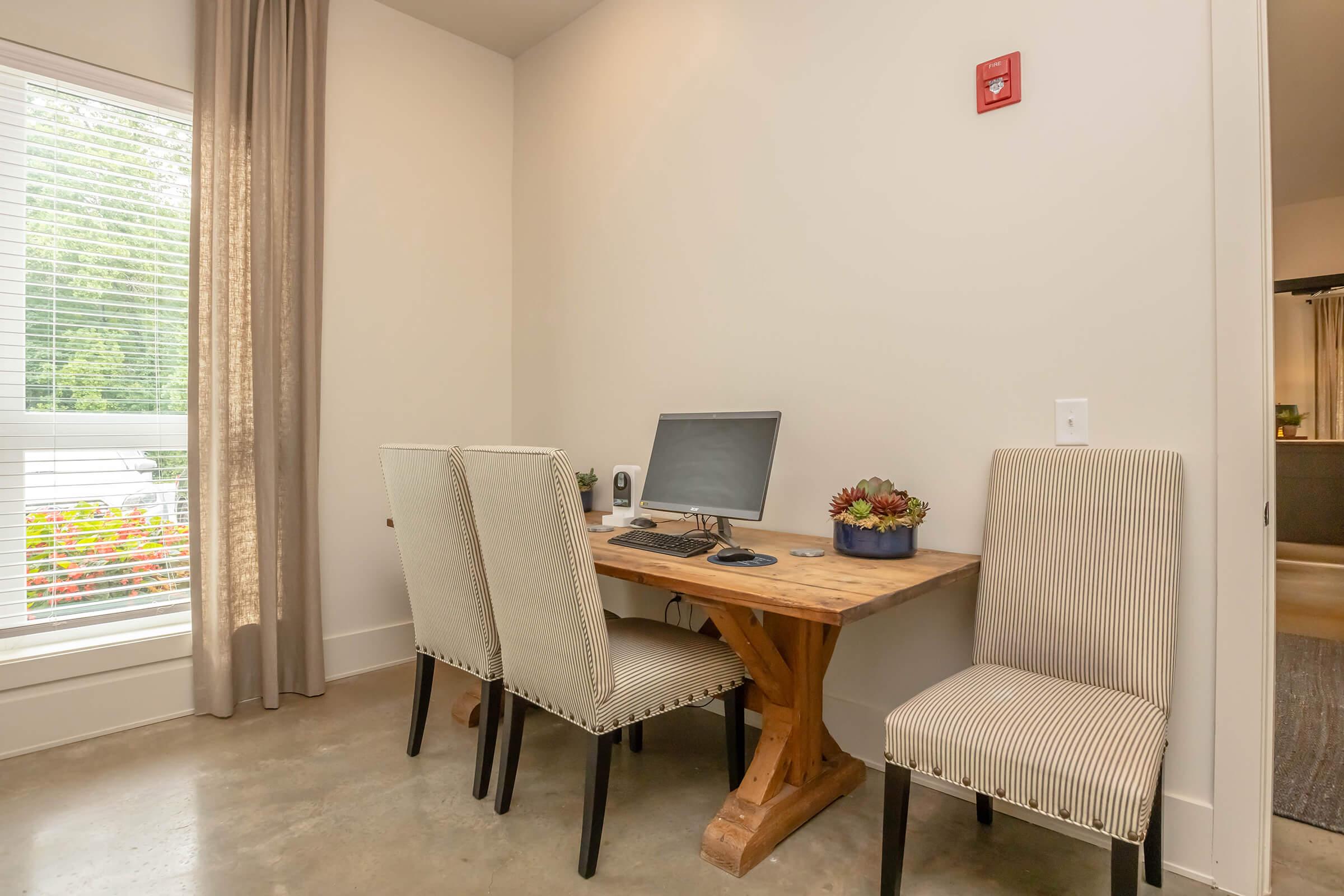 A small, minimalistic workspace featuring a wooden desk with a computer, two striped upholstered chairs, and a decorative plant. Natural light streams in through a window with sheer curtains, illuminating the room. The floor is a polished concrete surface, adding a modern touch to the decor.