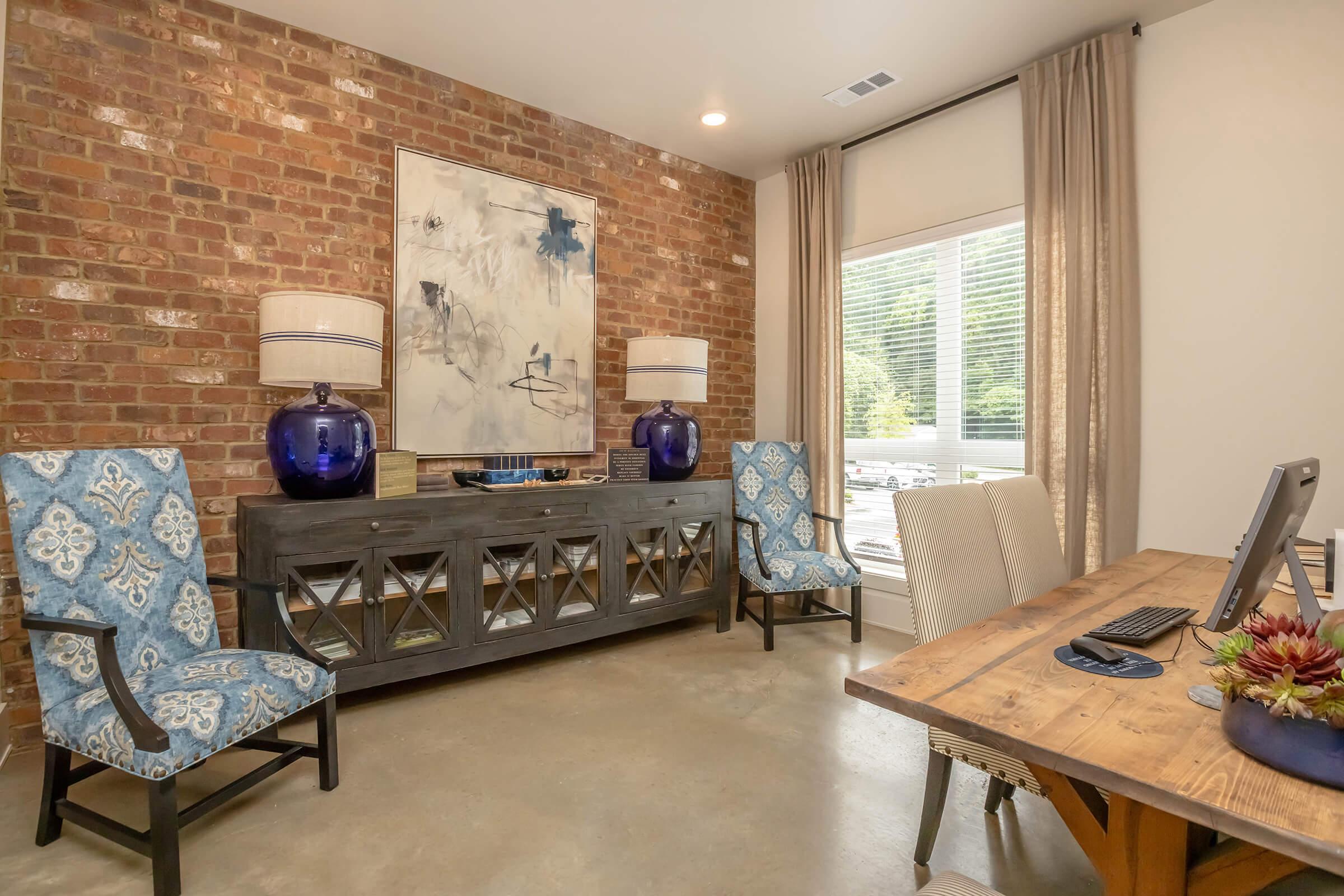 A modern home office featuring a brick accent wall, two upholstered blue chairs with white patterns, a dark wooden console table with decorative items, and a wooden desk with a computer. Large windows allow natural light to brighten the space, complemented by beige curtains.