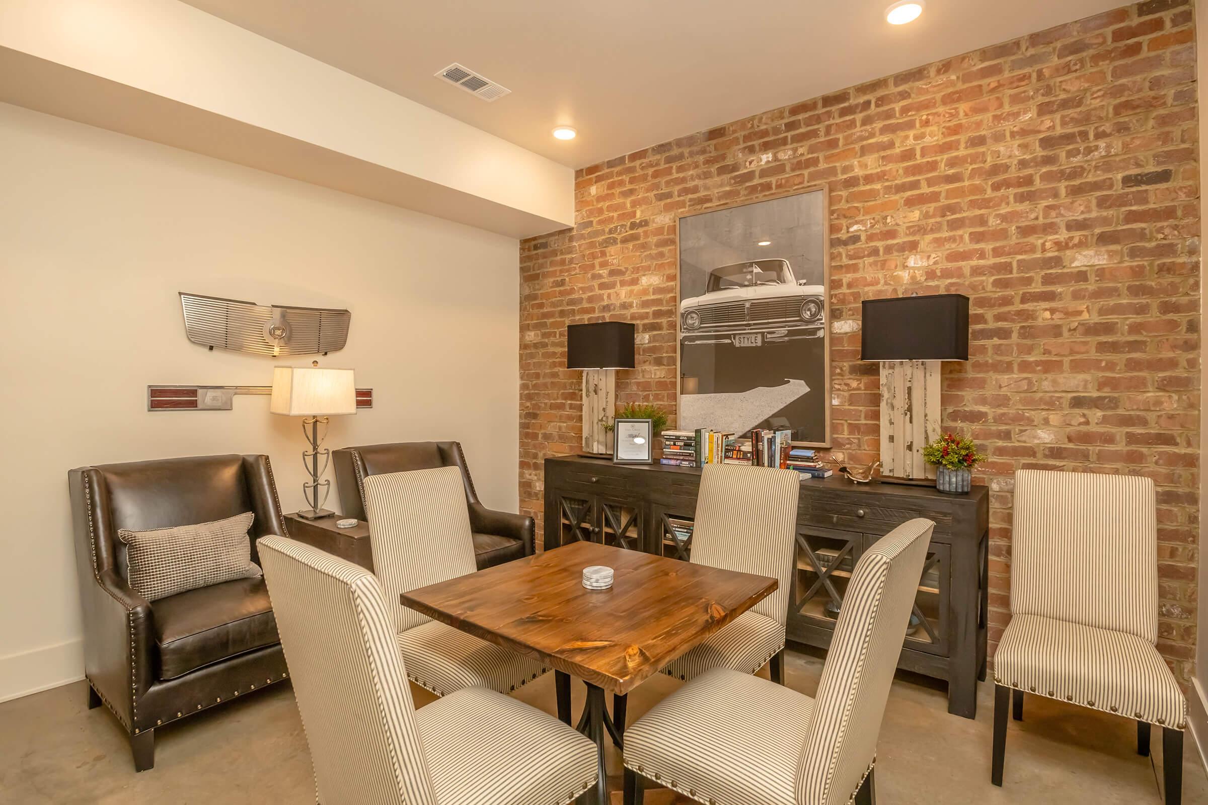 Modern living space featuring a wooden dining table surrounded by upholstered chairs. A cozy sitting area with leather armchairs is present, alongside a stylish sideboard. The room is accented by a brick wall and illuminated by warm lighting, with a vintage car-themed artwork above the sideboard.