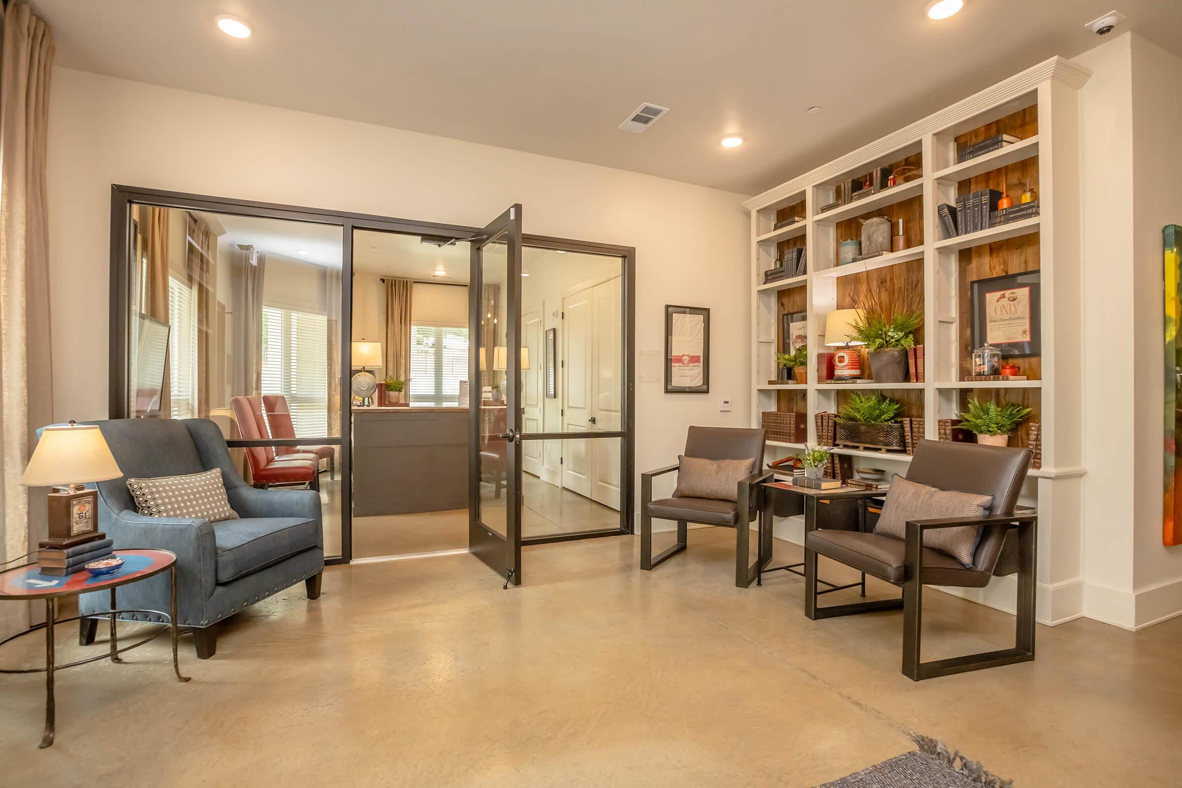 A modern office space featuring a glass entrance, stylish seating area with blue and gray chairs, a small table with a lamp, and a bookshelf adorned with plants and decorative items. Bright natural light filters through large windows, creating an inviting atmosphere.