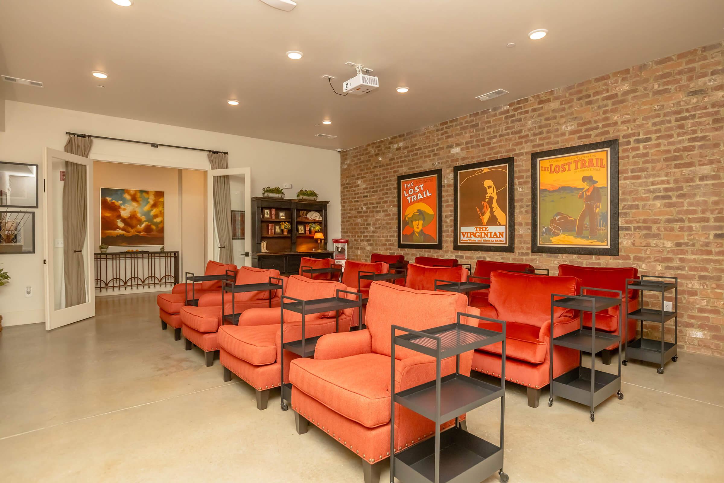 A cozy room with brick walls featuring vintage posters. It has several orange armchairs arranged in rows, accompanied by small black carts. A projector is mounted on the ceiling, and there is a doorway leading to another space in the background, enhancing the inviting atmosphere.