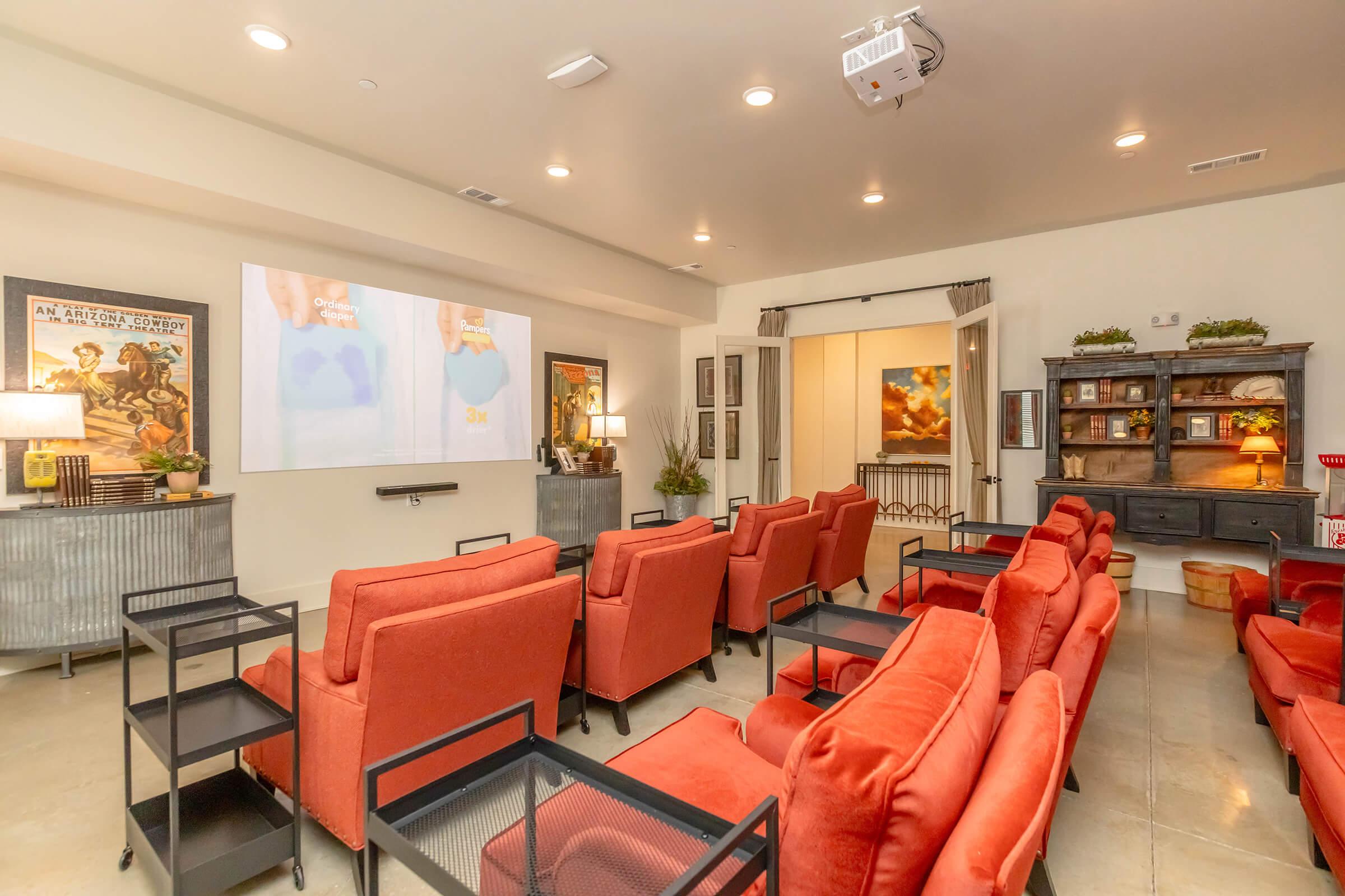 A modern lounge area featuring several red armchairs with side tables arranged in a theater-style layout. A large screen is mounted on the wall, displaying an image. The room is well-lit with neutral colors and artwork adorning the walls, creating a cozy and inviting atmosphere.