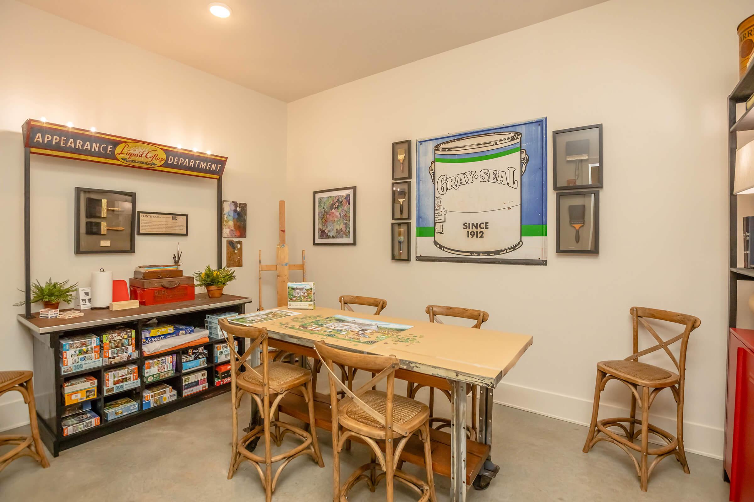 A bright, inviting room featuring a large table surrounded by wooden chairs. On the walls, there are framed artworks, including a vintage-style poster. A shelf displays various boxes and decorative items. The overall atmosphere is creative and welcoming, suggesting a space for art or group activities.