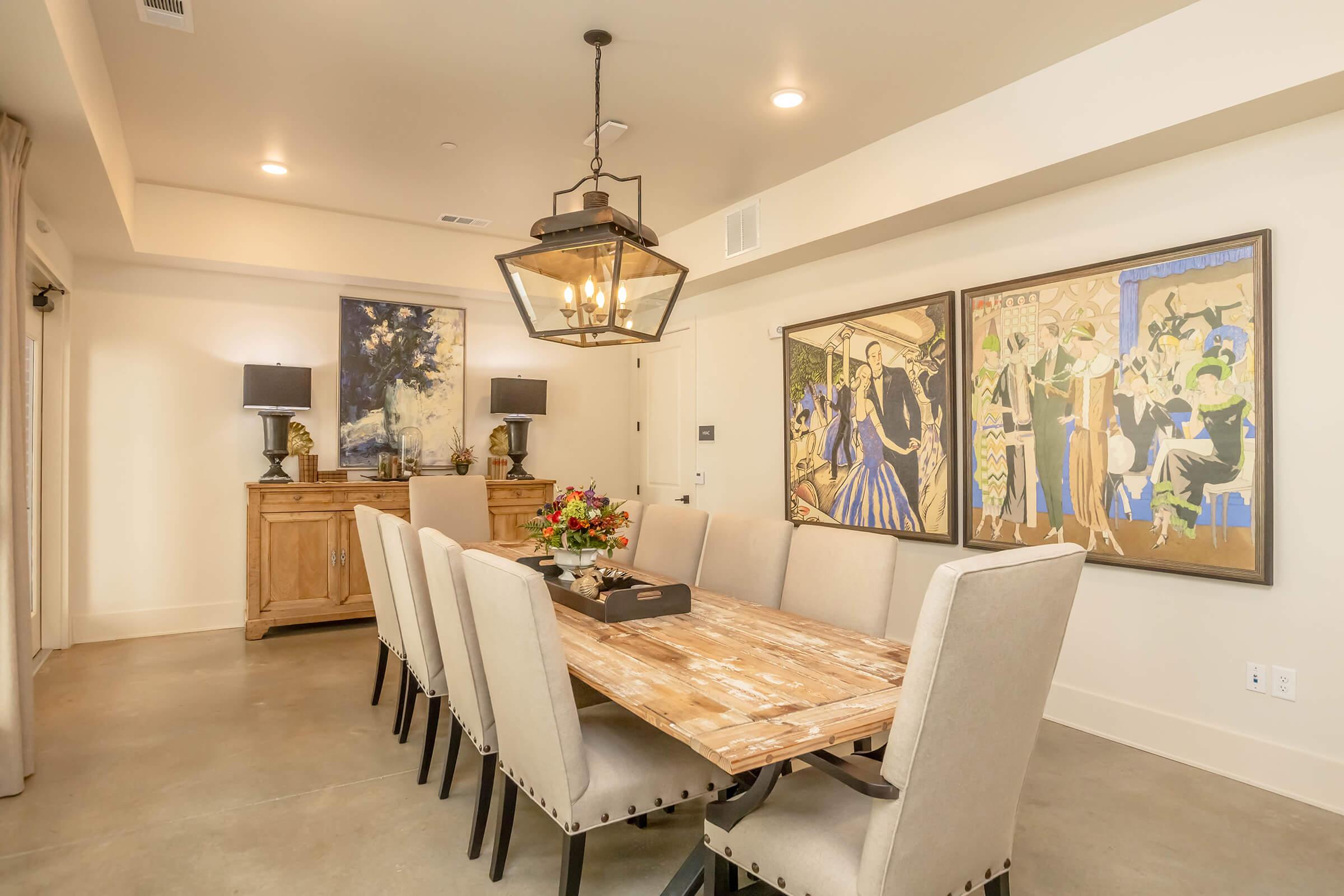 A modern dining room with a large wooden table surrounded by beige upholstered chairs. The walls feature two large framed artworks, and there are two lamps on a wooden sideboard. A bouquet of flowers sits in the center of the table, and the space is well-lit with a pendant light.
