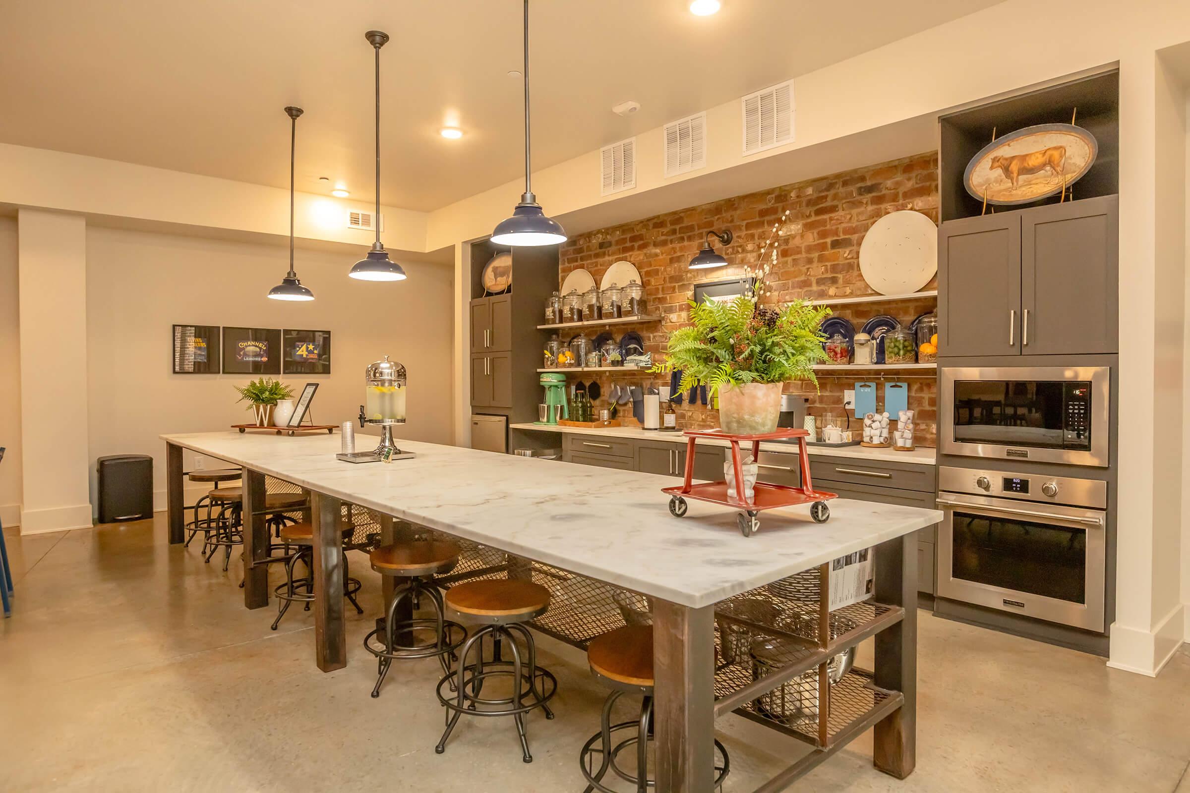 A modern kitchen featuring a large marble island with bar stools, industrial-style lighting, and an exposed brick wall. The space includes open shelving with decorative plates and plants, a built-in oven, and a cozy dining area, creating a warm and inviting atmosphere.