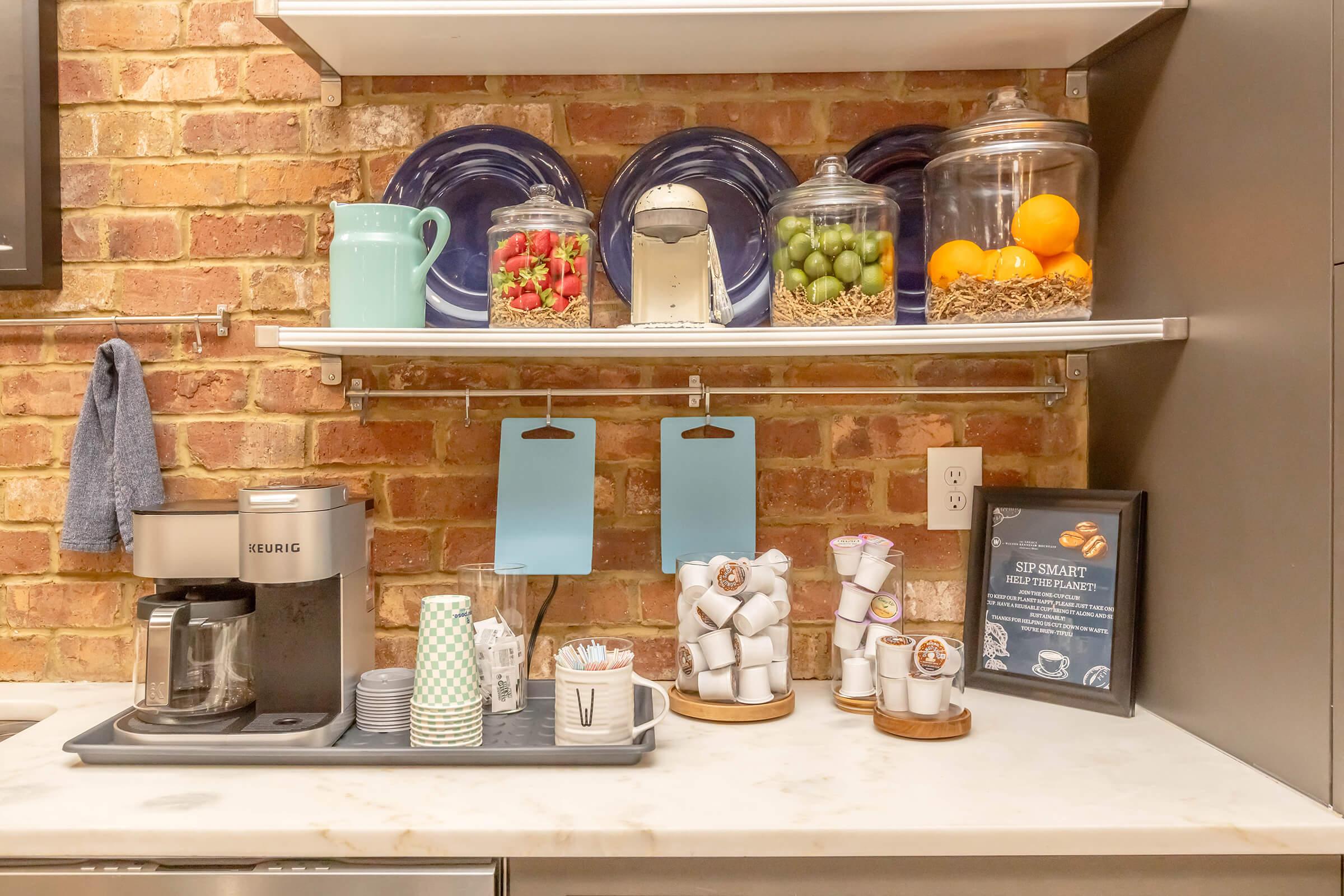 A modern kitchen shelf featuring a coffee maker, colorful ceramic cups, jars filled with fruits and snacks, and a collection of white coffee pods. Behind, brick walls add a rustic charm, while two blue notepads hang from the shelf for notes or reminders.