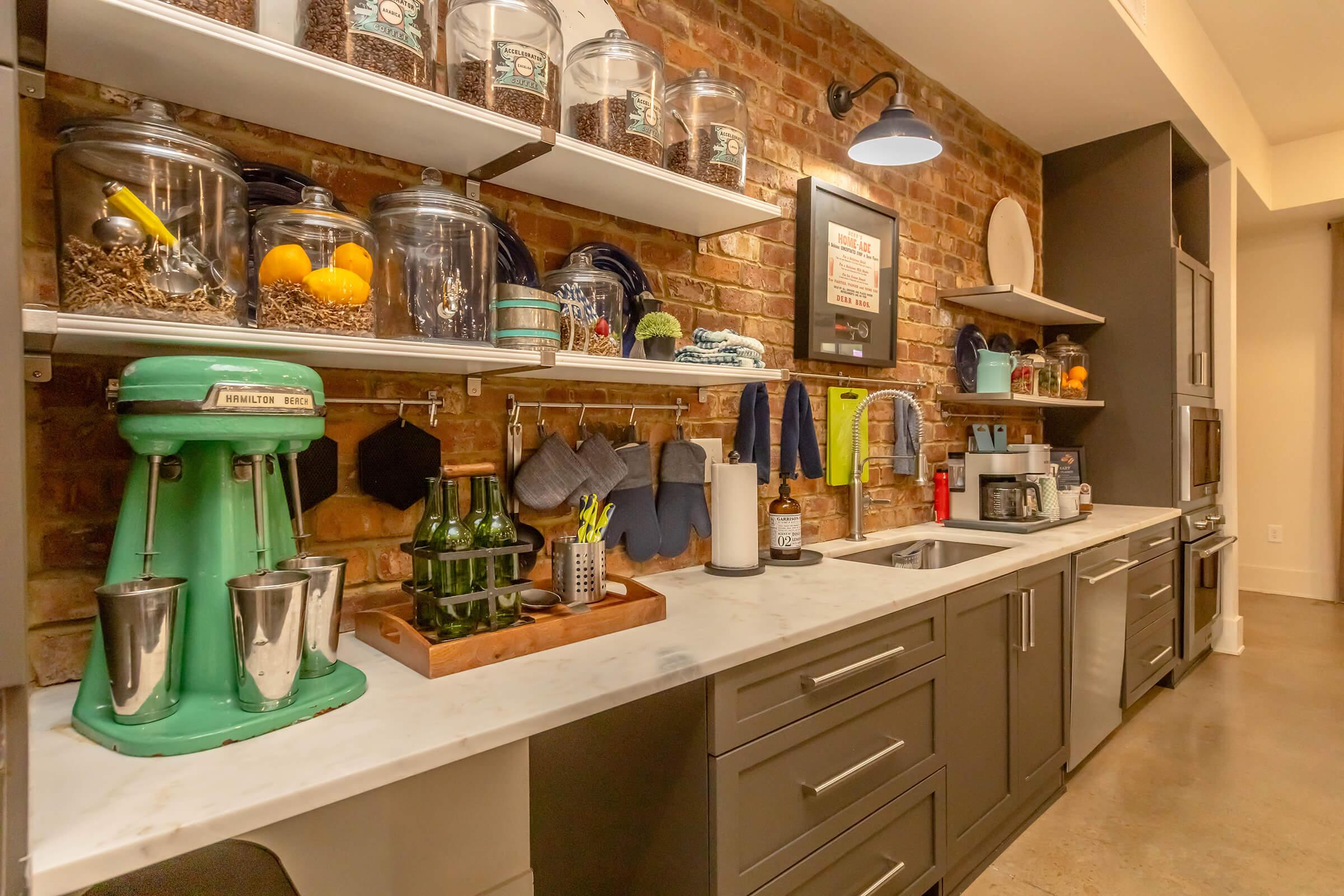 A stylish kitchen featuring a mix of modern and vintage elements. There are shelves displaying glass jars with food items, a mint green mixer, a wooden cutting board, and various kitchen utensils. The backdrop includes exposed brick walls and gray cabinets, creating a warm and inviting atmosphere.