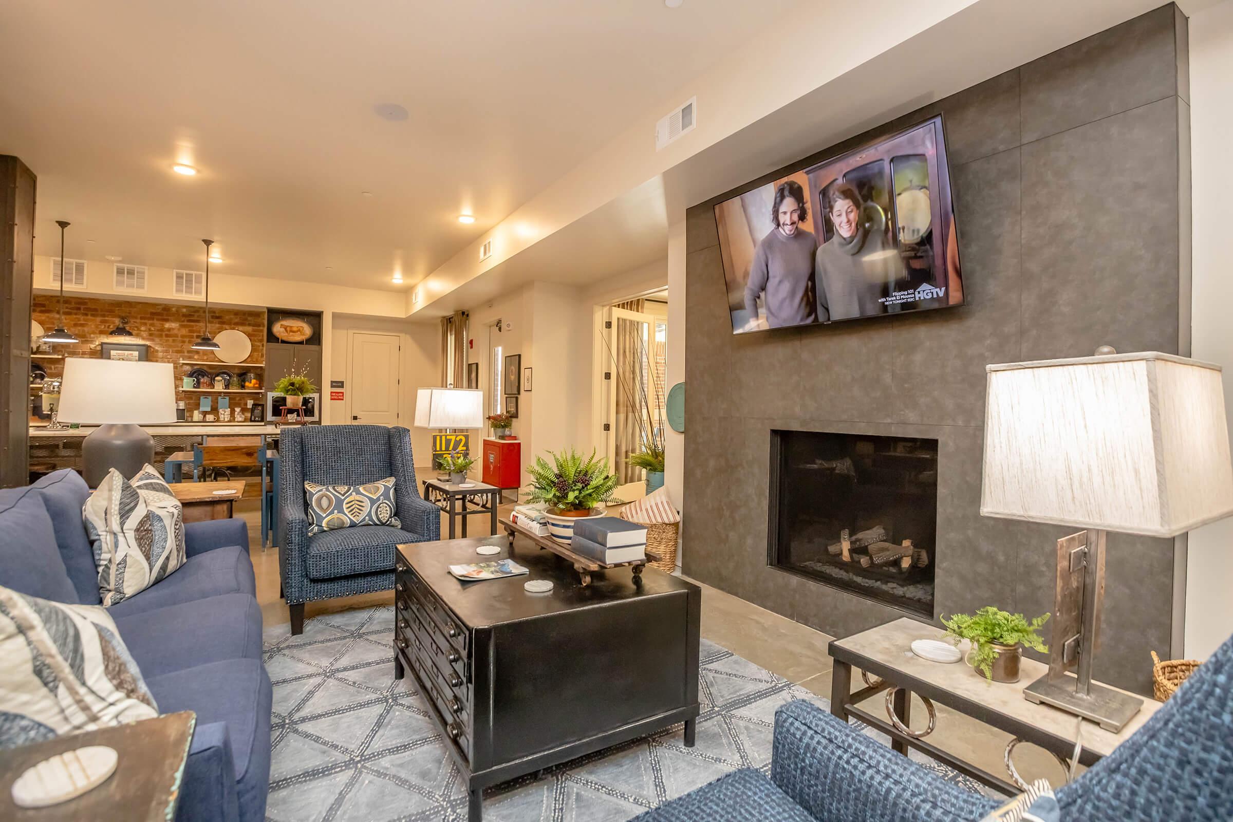 A cozy living room featuring a blue sofa, patterned throw pillows, and a coffee table with decorations. There's a wall-mounted TV displaying a show, and a fireplace beneath it. The room has warm lighting, a textured rug, and a small plant on the coffee table, creating an inviting atmosphere.