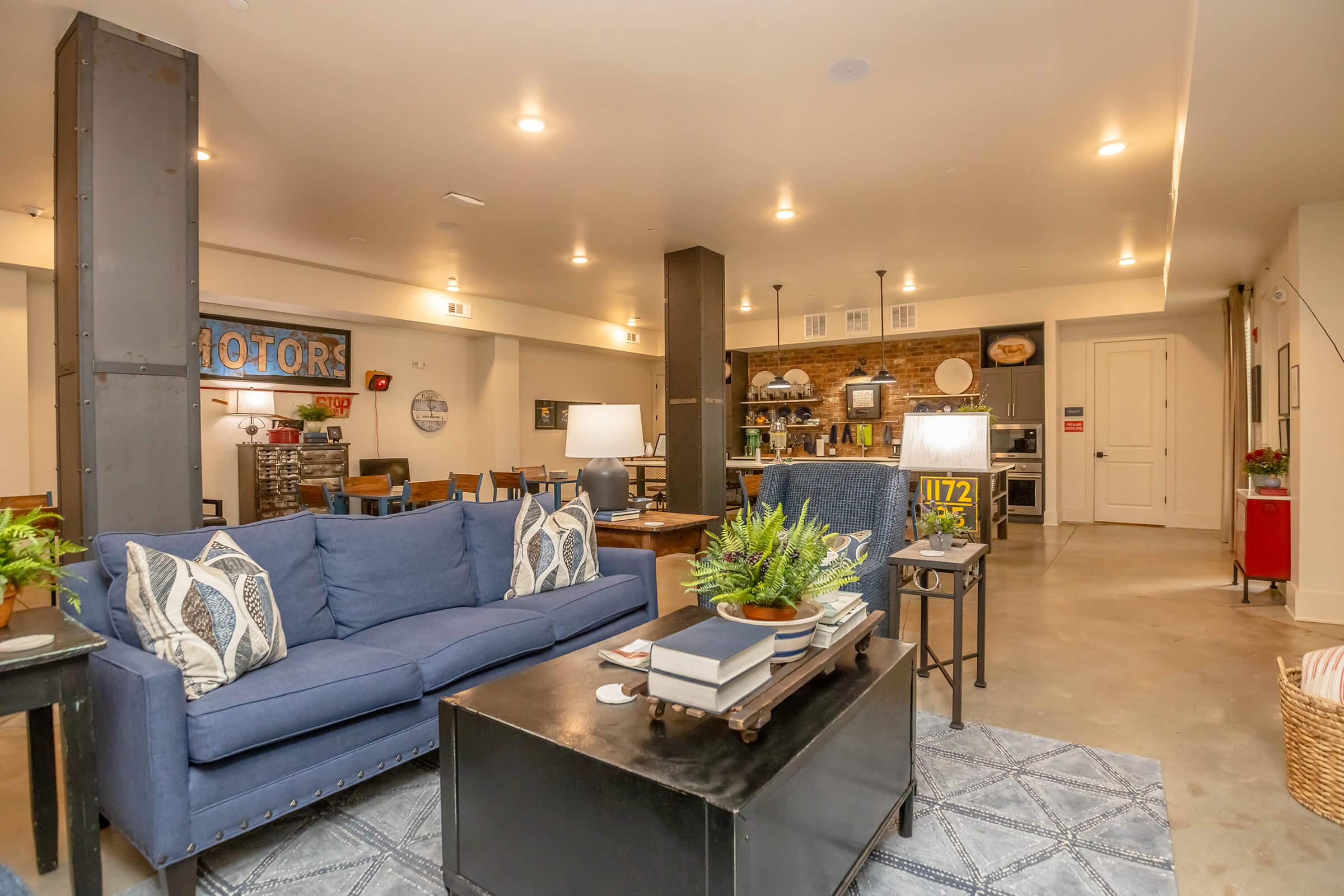 A spacious modern living area featuring a cozy blue sofa with decorative pillows, a stylish coffee table, and a decorative rug. In the background, there's a dining area with wooden tables and chairs, a well-equipped kitchen with stone accents, and various decorative items on display. Warm lighting enhances the inviting atmosphere.