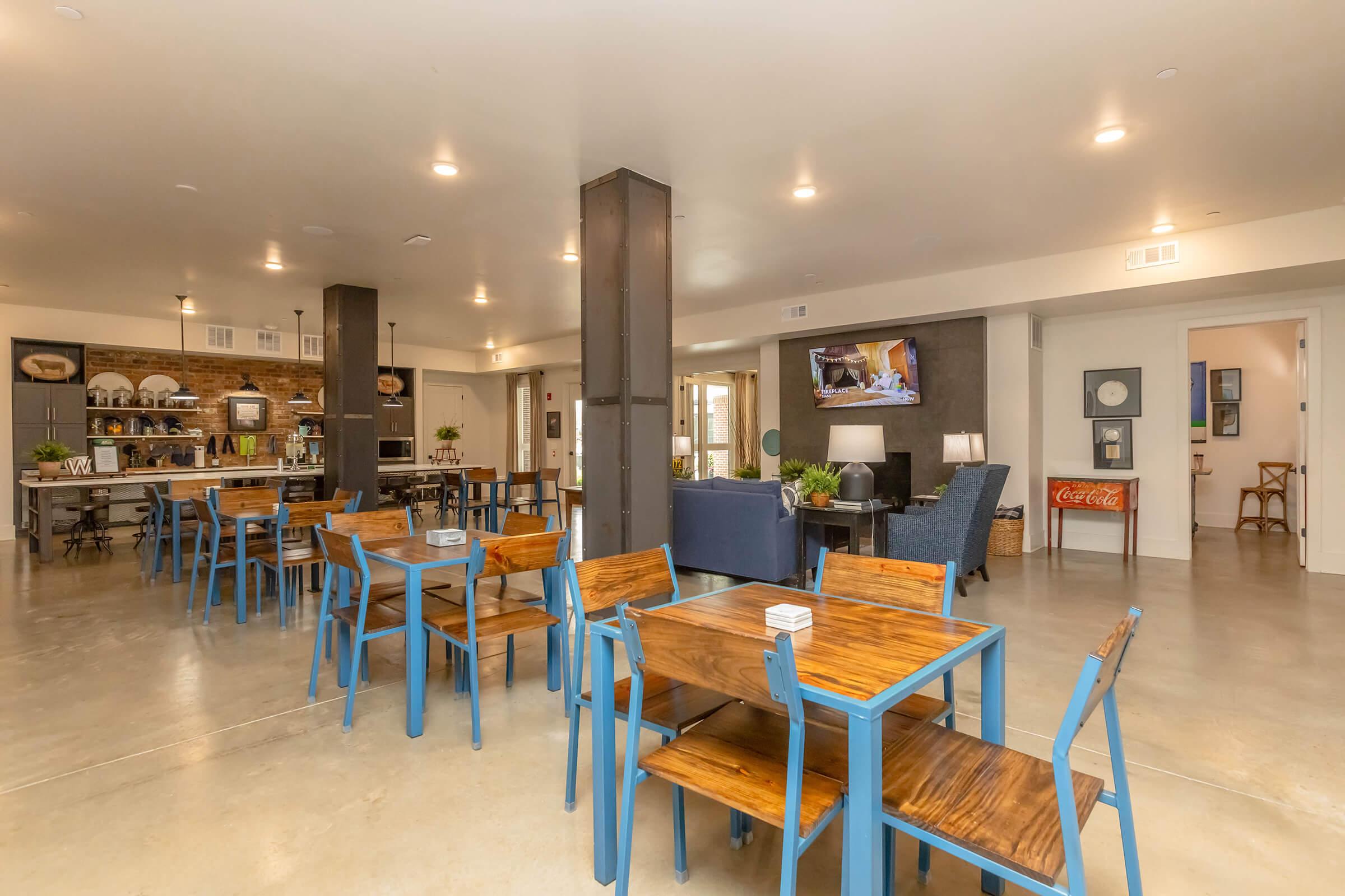 A modern, spacious communal area featuring blue wooden tables and chairs. The room includes a kitchen area with shelves filled with dishes, a television mounted on the wall, and seating in the corner. Bright lighting creates a welcoming atmosphere with polished concrete floors.