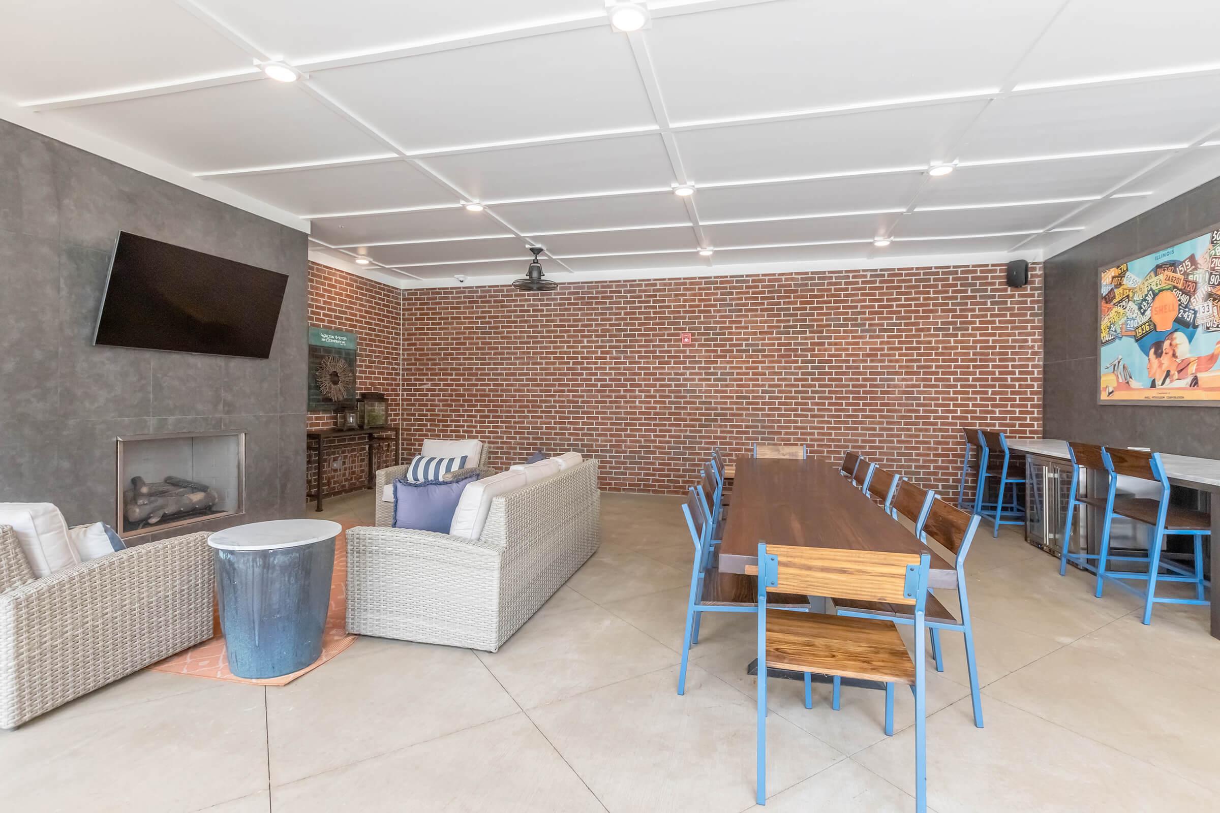 Modern communal space featuring a long wooden dining table with blue chairs, a comfortable sofa, and a television mounted on a gray wall. The exposed brick background adds a rustic touch, complemented by a decorative piece on the wall. The room has an airy feel with ample lighting.