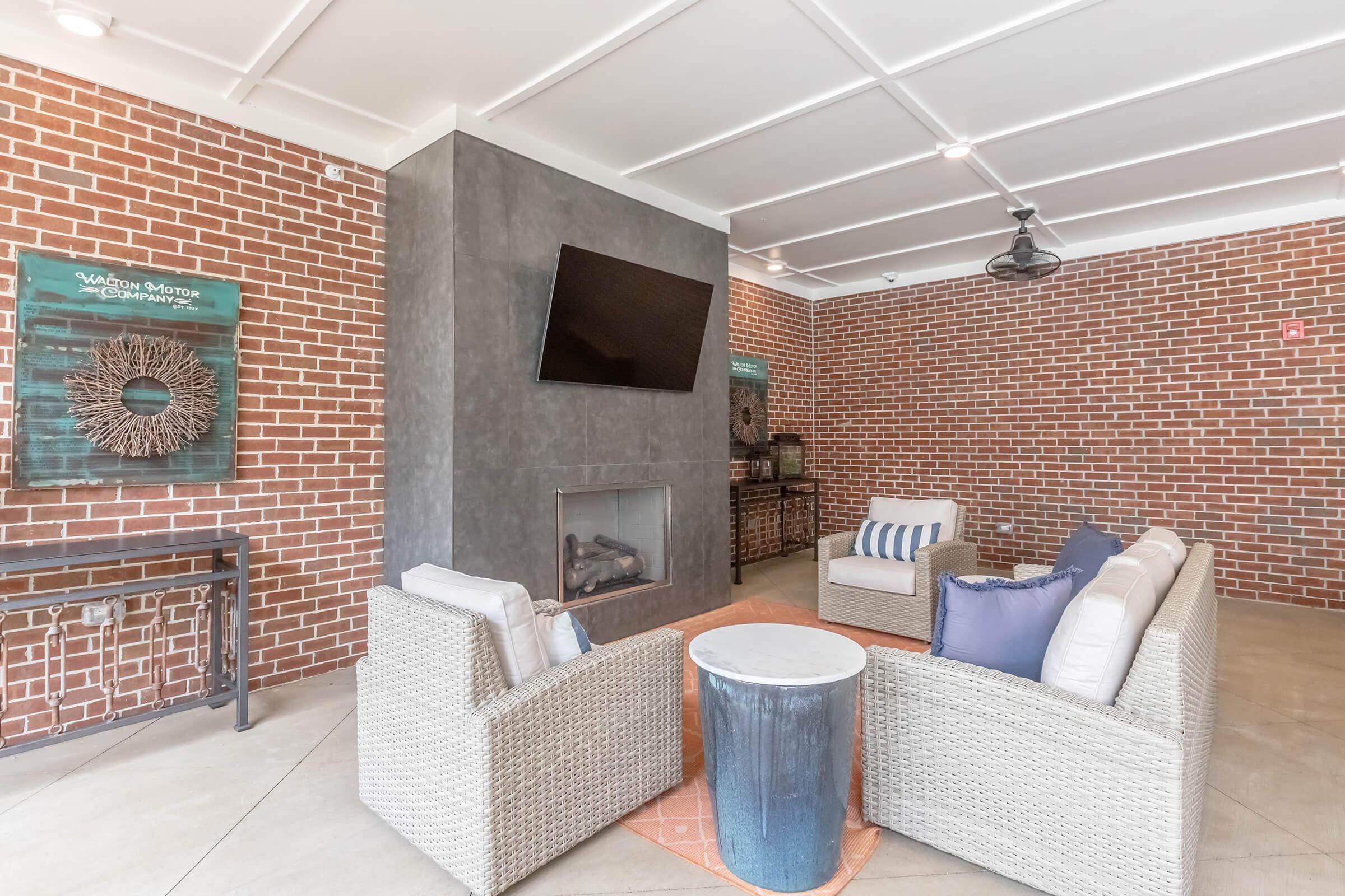 A cozy lounge area with a modern design featuring a brick wall, a flat-screen TV mounted above a fireplace, and comfortable seating made of light-colored fabric. There is a small round table and decorative wall art, creating an inviting atmosphere for relaxation or socializing.