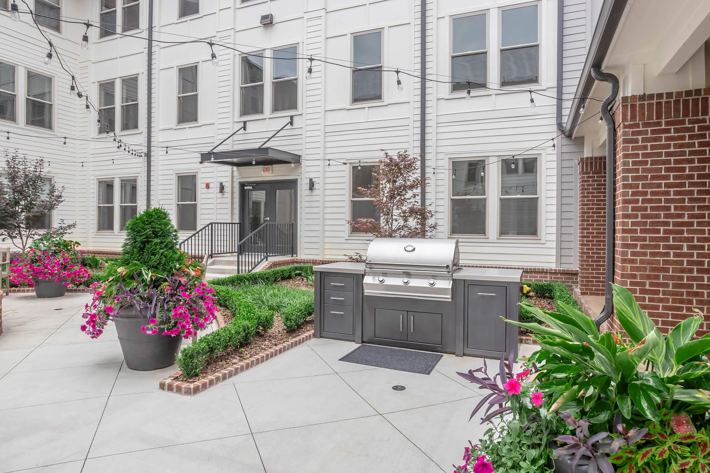 An outdoor BBQ grill is situated in a landscaped courtyard surrounded by vibrant flower beds. The area features white buildings with large windows and string lights overhead, creating an inviting atmosphere for gatherings and outdoor cooking.