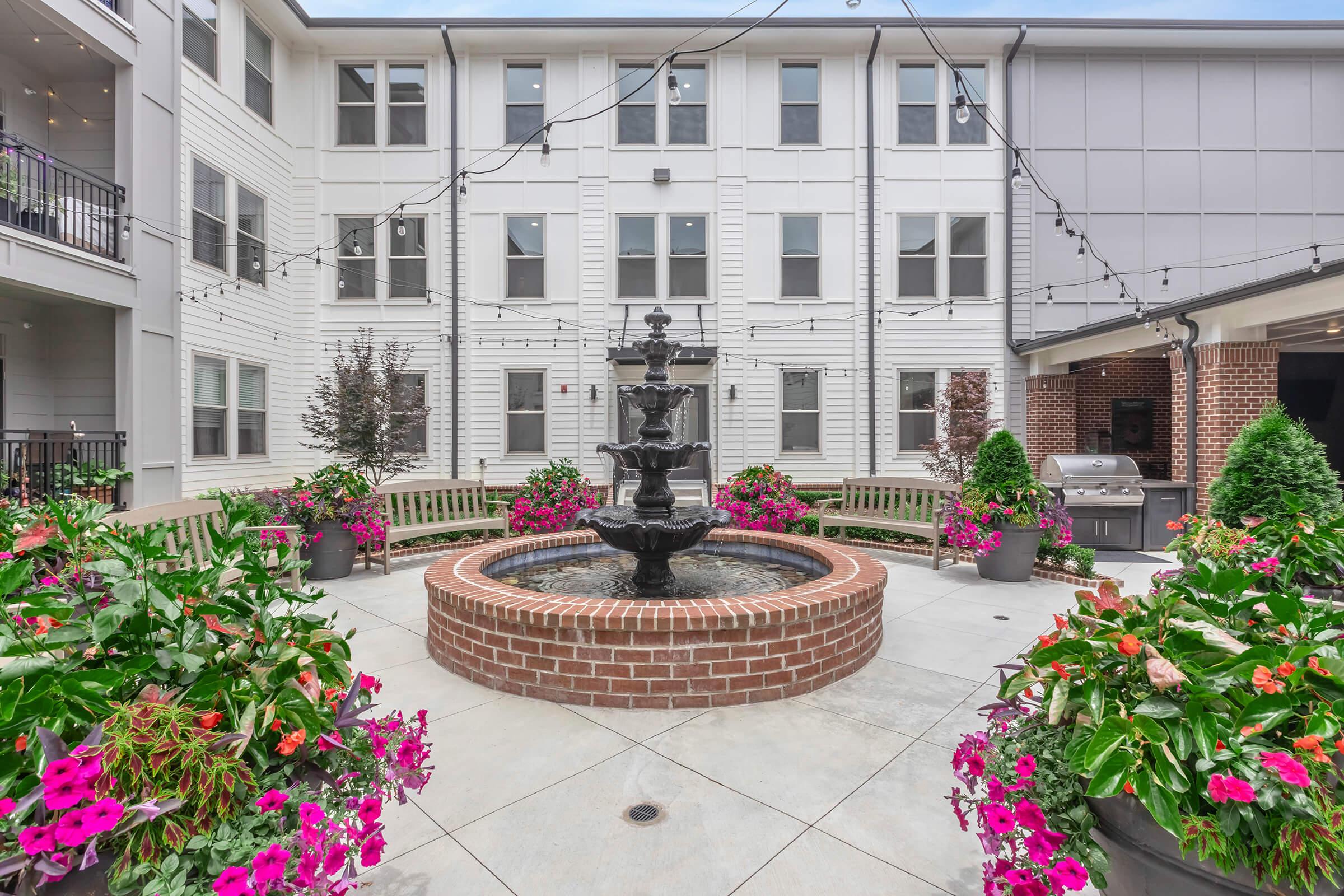 A serene courtyard featuring a decorative fountain at the center, surrounded by vibrant flowers in planters. Benches encircle the fountain, creating a cozy seating area. Strings of warm lights hang overhead, adding ambiance to the outdoor space. Nearby, a grill is visible, inviting gatherings and relaxation.