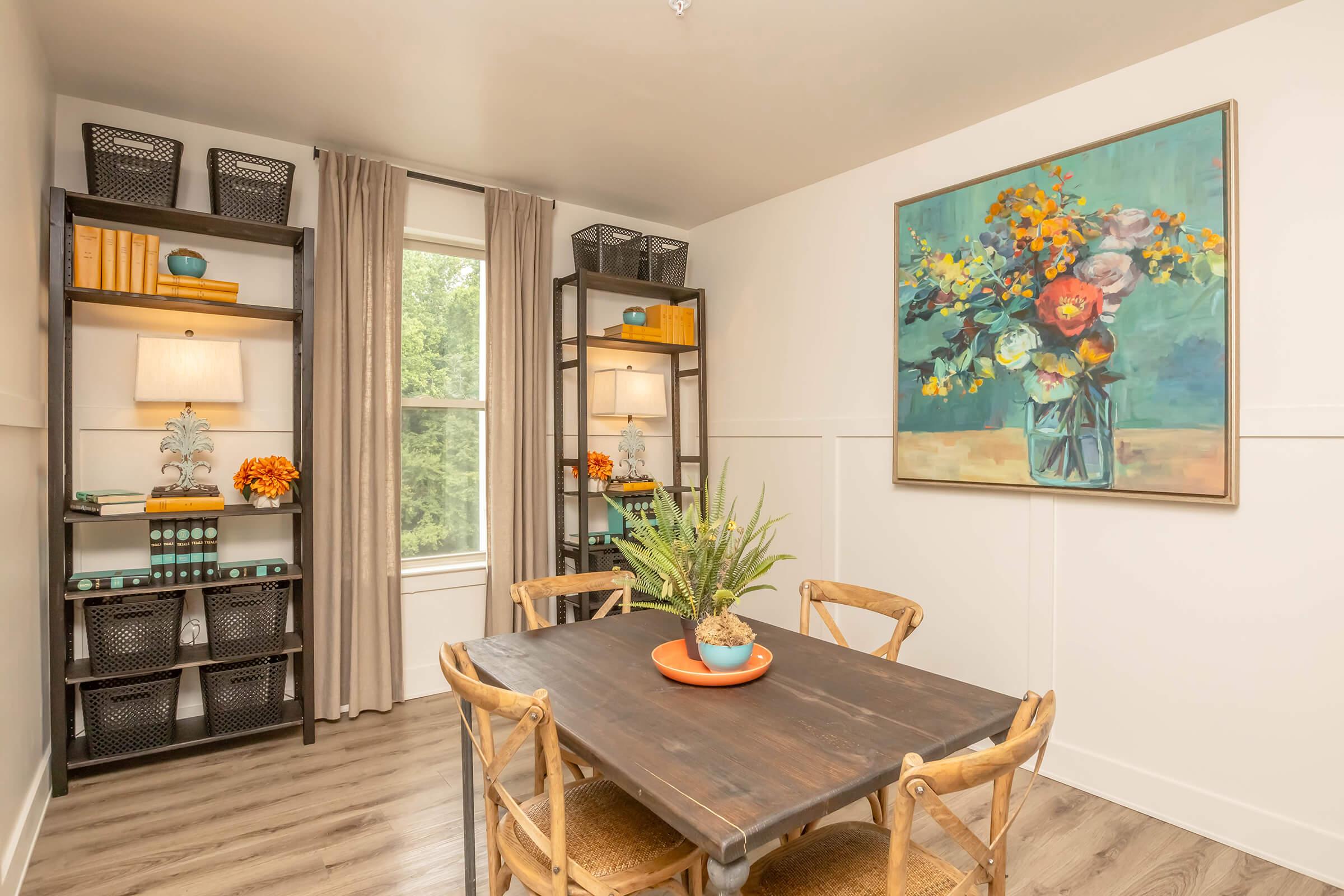 A cozy dining area featuring a wooden table surrounded by chairs. On the walls, there are two decorative shelves with books and small plants. A large floral painting hangs above the table, and natural light enters through a window with sheer curtains. The décor combines a modern and welcoming aesthetic.