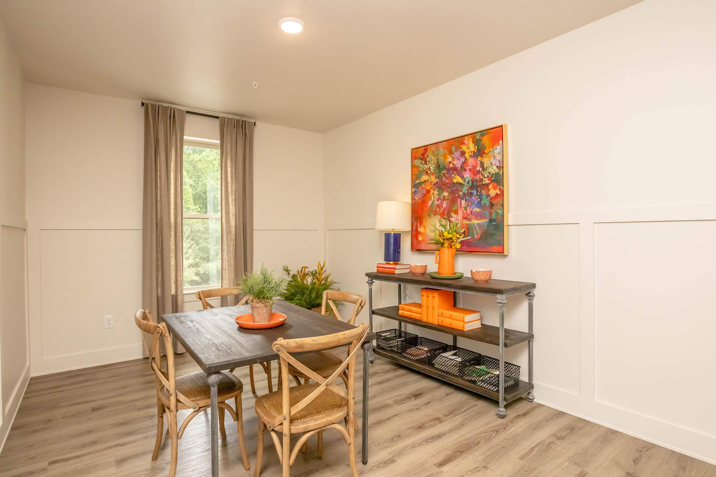A cozy dining area featuring a wooden table with chairs, a colorful artwork on the wall, and a side shelf displaying orange accents. Natural light comes in from a large window covered with sheer curtains, and potted plants add a touch of greenery to the inviting space.