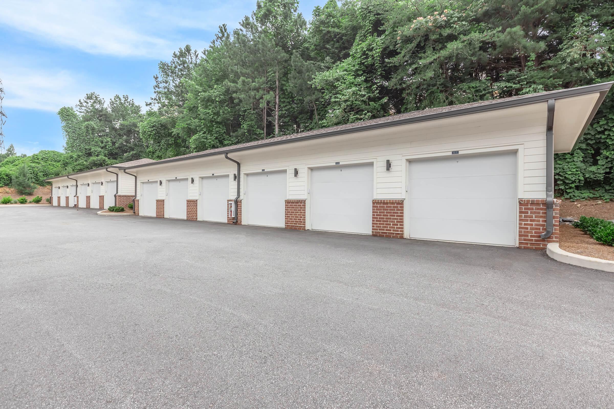 Row of closed garage doors with a paved driveway in front, surrounded by greenery and trees. The garages have brick accents and are part of a larger building structure, set in a tranquil outdoor setting. The sky is clear and blue, enhancing the peaceful atmosphere.