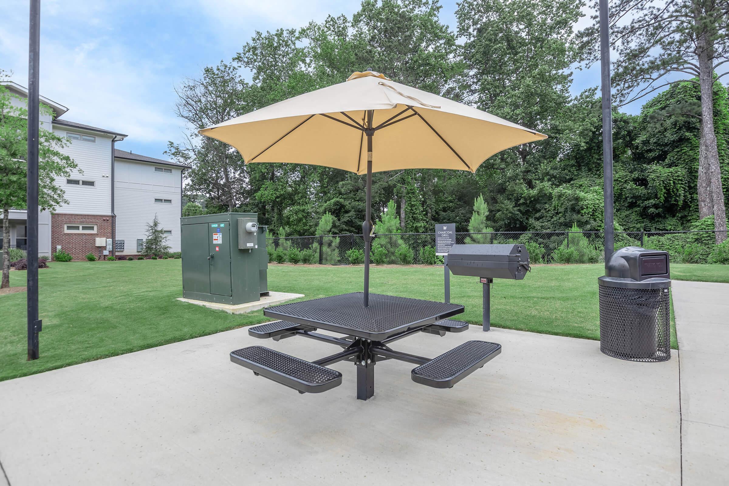 A picnic table with a large umbrella sits on a concrete surface, surrounded by green grass and trees. Nearby, there's a grill, a trash can, and a utility box, creating a recreational space for outdoor gatherings.