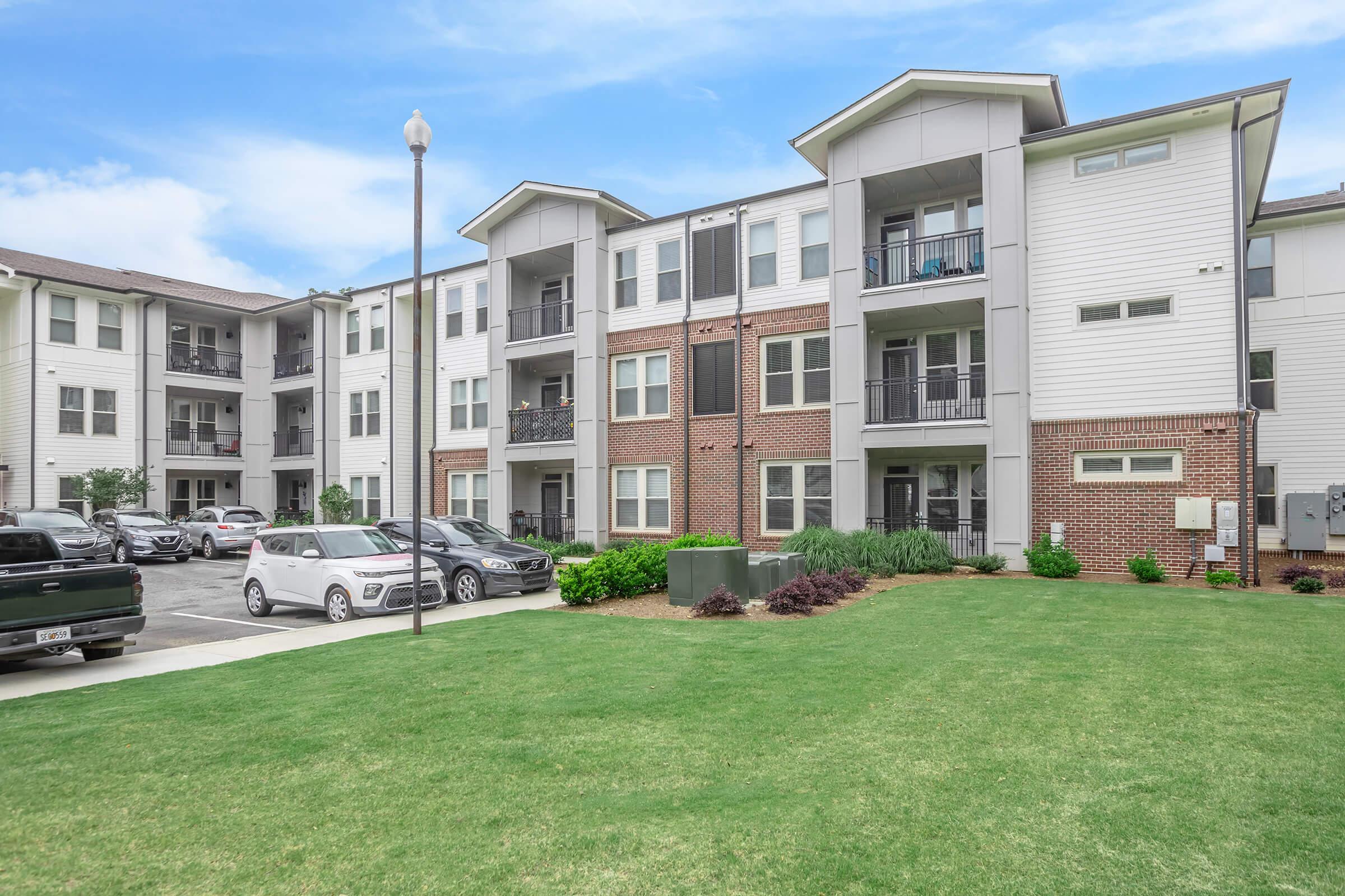 A modern apartment building with multiple floors, featuring a mix of brick and siding exteriors. The landscaped lawn in front includes green grass and shrubs. Several parked cars are visible in the foreground, and the sky is partly cloudy.