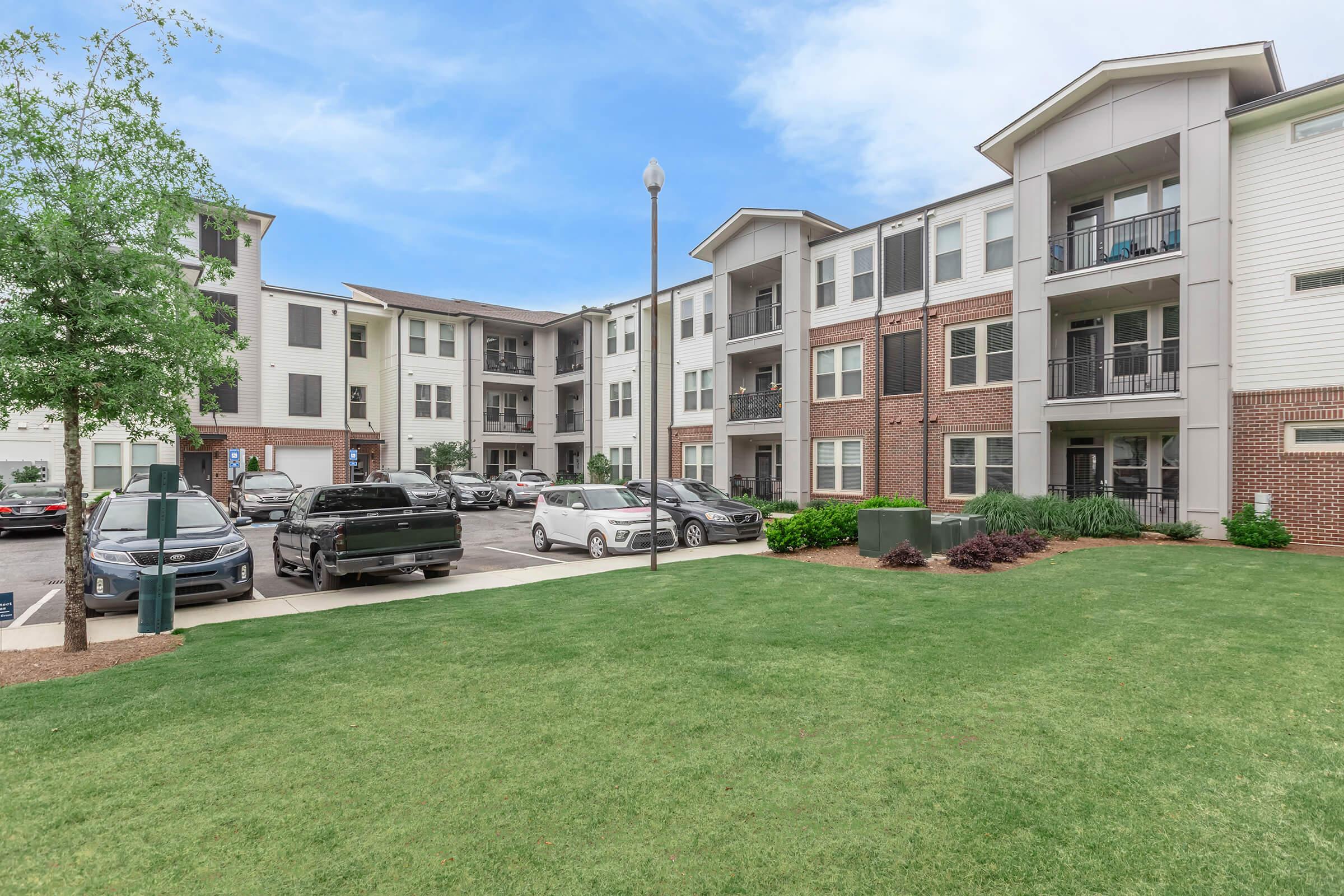 A view of a modern apartment complex featuring multiple buildings with a mix of brick and siding. Lush green grass is in the foreground, with parked cars in a lot adjacent to the buildings. Trees are visible, adding to the landscaped surroundings. Clear blue sky overhead.