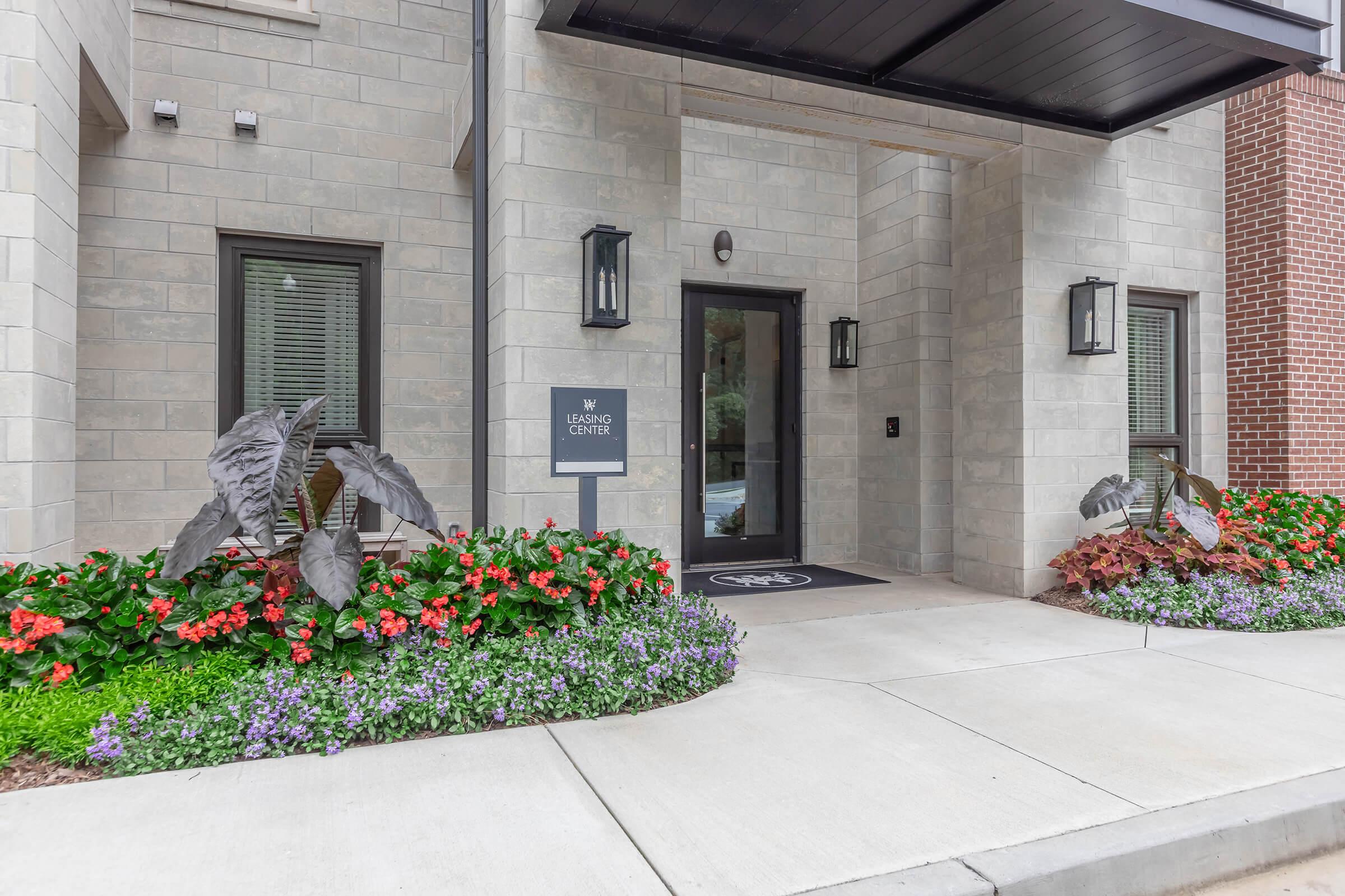 A modern building entrance featuring a dark door with glass panels, flanked by two lanterns. The pathway is bordered by a colorful flowerbed with red, purple, and white blooms. A sign labeled "Nursing Center" is mounted on the wall beside the entrance. The facade is composed of light gray stone.