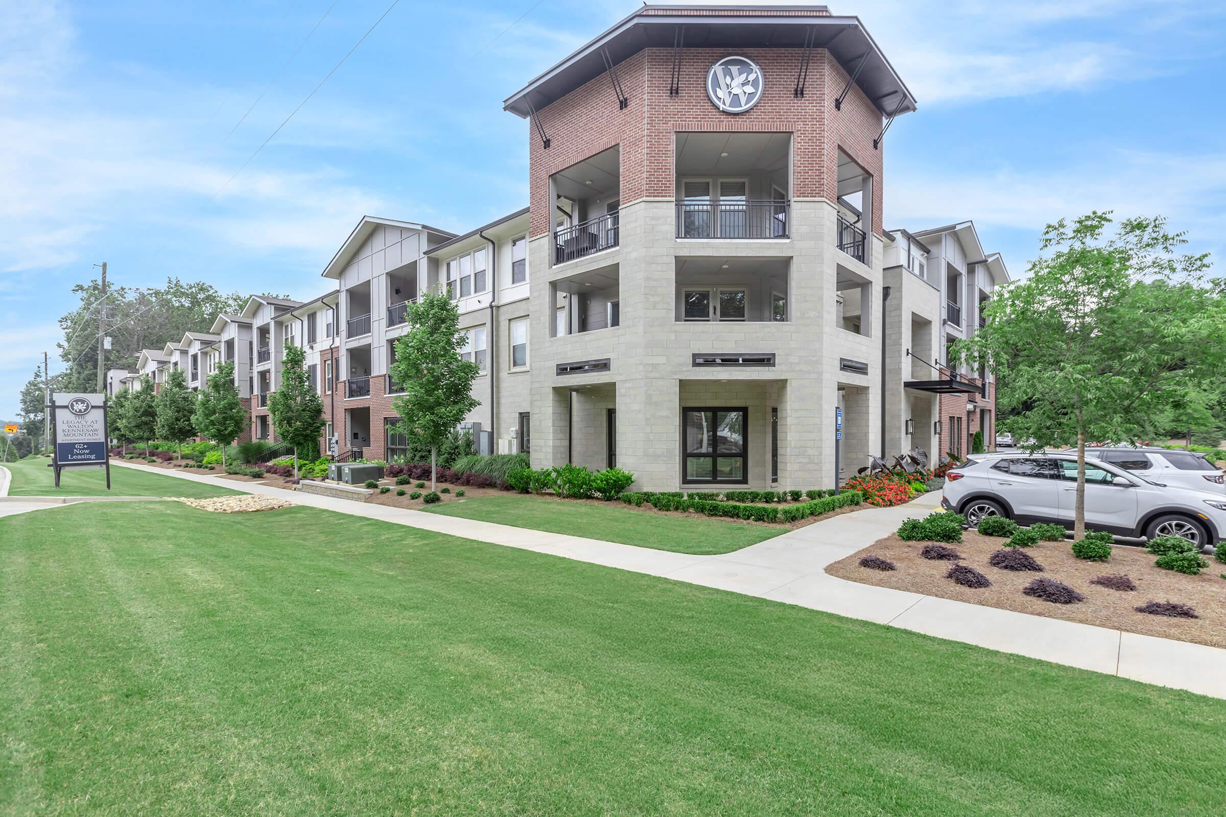 A modern apartment complex featuring a two-story building with brick accents and large balconies, surrounded by landscaped green lawns and decorative plants. A pathway leads through the property, with a sign visible at the entrance and a parked car nearby under a clear blue sky.