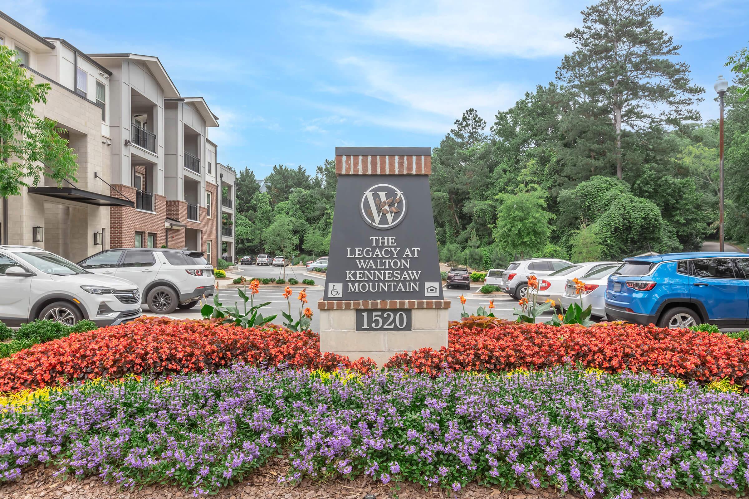 A landscaped entrance sign for "The Legacy at Walton Kennesaw Mountain" featuring colorful flowers and greenery. The sign displays the name and address "1520" with modern brick and stone accents, surrounded by parked cars in a well-maintained area.