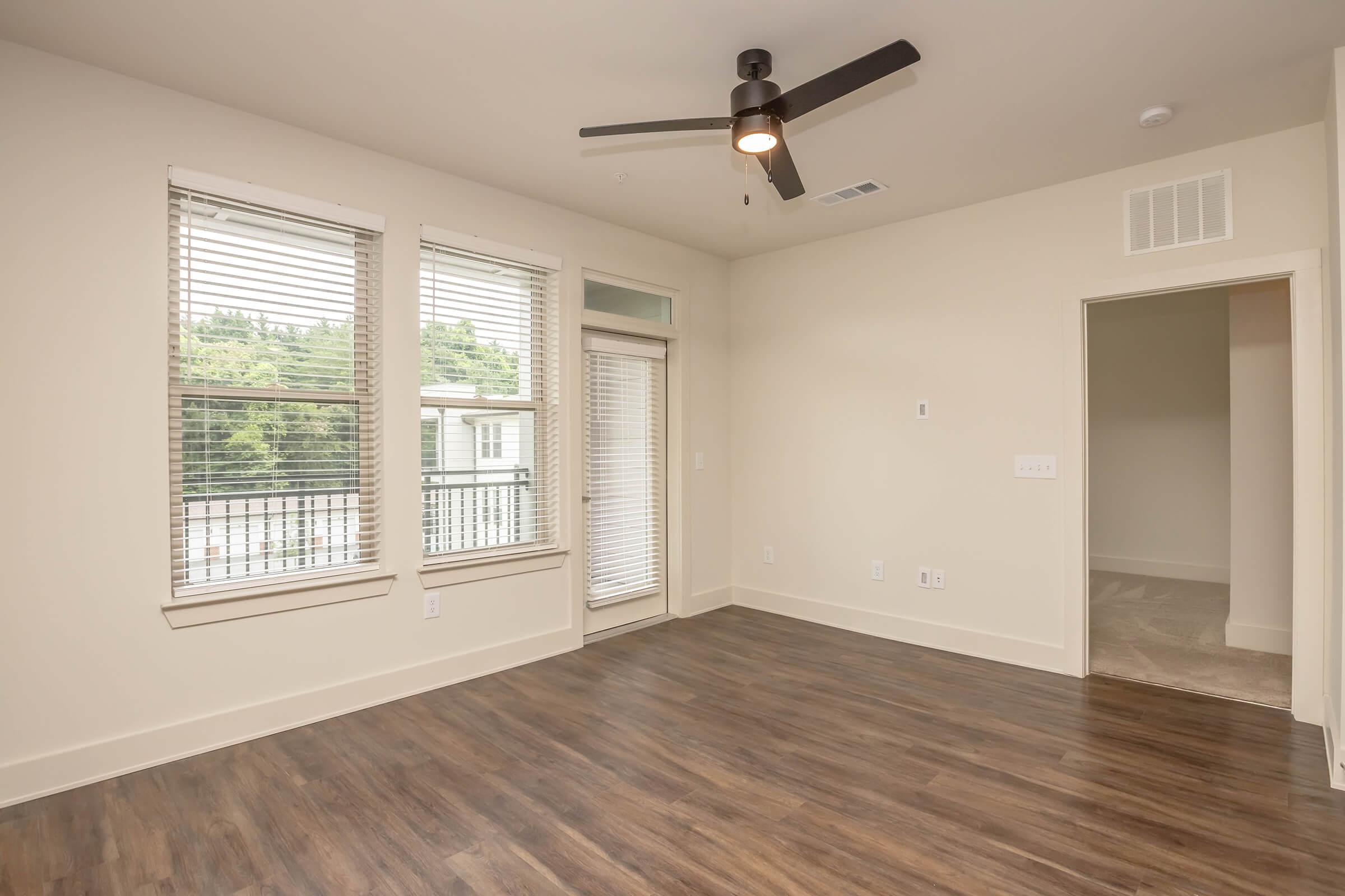 A bright, empty living room featuring large windows with blinds, a ceiling fan, and wooden flooring. There’s a sliding door leading to a balcony, and a doorway on the right side leading to another room. The walls are painted in a light color, creating an open and airy feel.