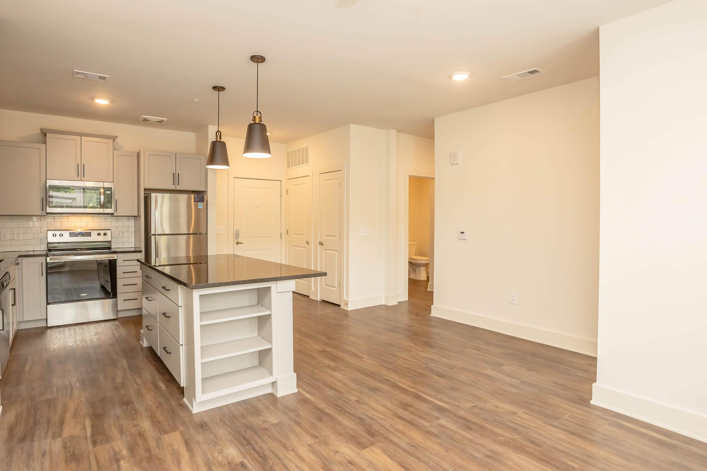 Modern kitchen featuring stainless steel appliances, a large island with seating, and light-colored cabinetry. The open layout includes a living area with hardwood floors and recessed lighting. A doorway leads to a bathroom in the background, with light walls enhancing the spacious feel.