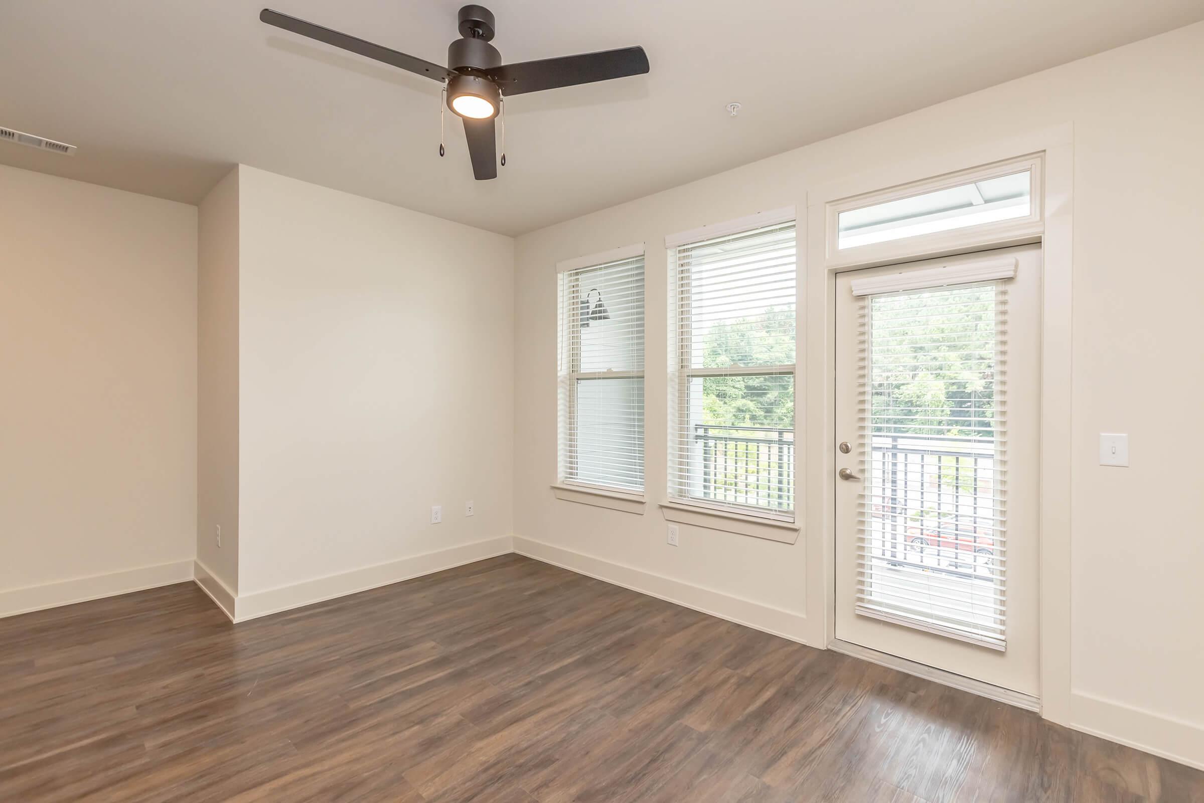 A spacious, well-lit living room featuring large windows with white blinds, a ceiling fan, and light-colored walls. The room has a wood-like laminate floor and a sliding glass door that leads to a small balcony. The overall ambiance is modern and inviting, suitable for relaxation or entertaining.