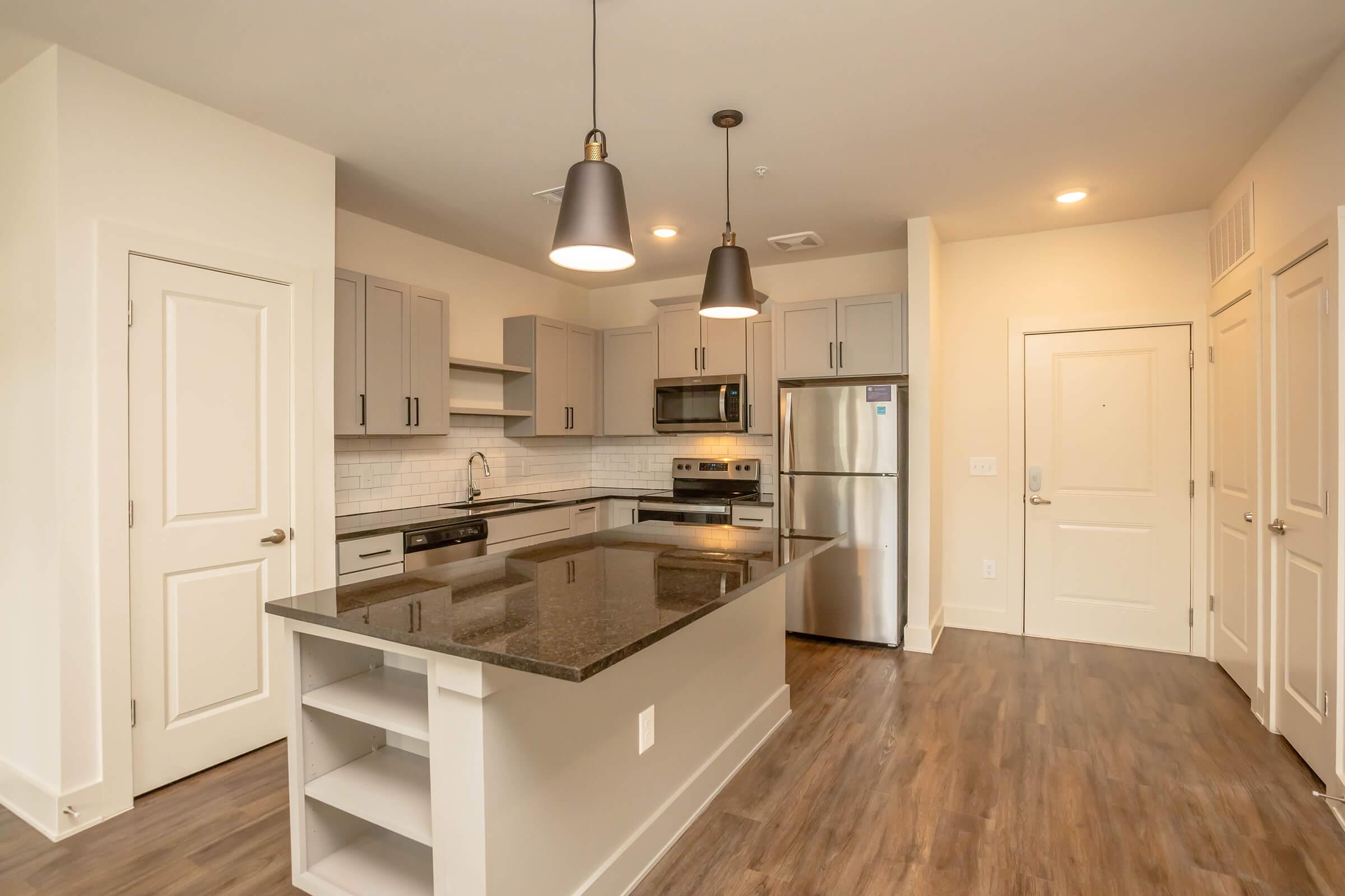 Modern kitchen with gray cabinetry, stainless steel appliances, and a large island with a dark countertop. Two pendant lights hang above the island, and there are white walls and wooden flooring. The room has an open layout, leading to a door and additional cabinetry on the side.