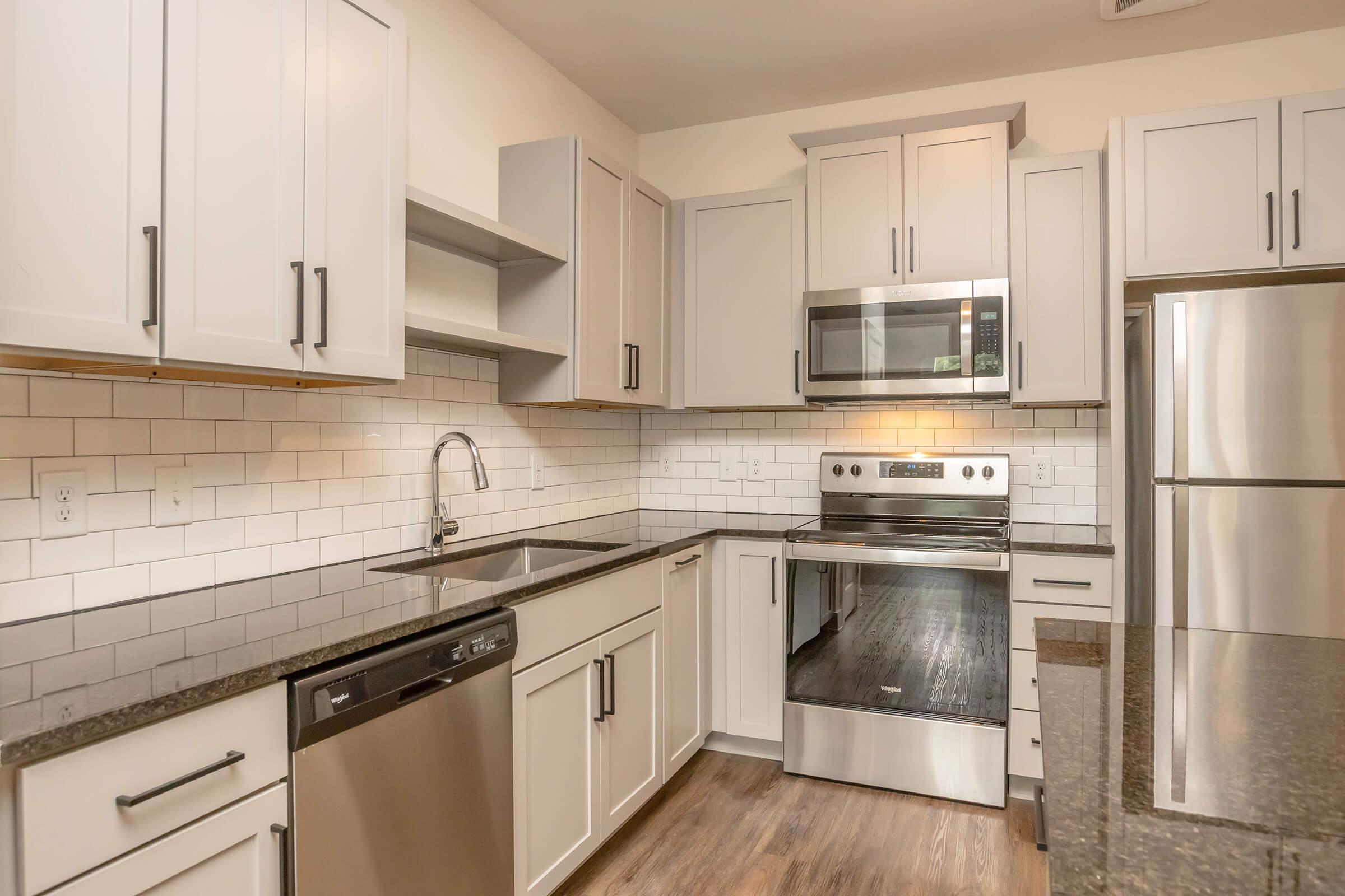 Modern kitchen featuring light gray cabinetry, stainless steel appliances including a microwave and oven, a dark granite countertop, and a tile backsplash with white subway tiles. The kitchen is well-lit, with a spacious design and a dishwasher integrated into the cabinetry.