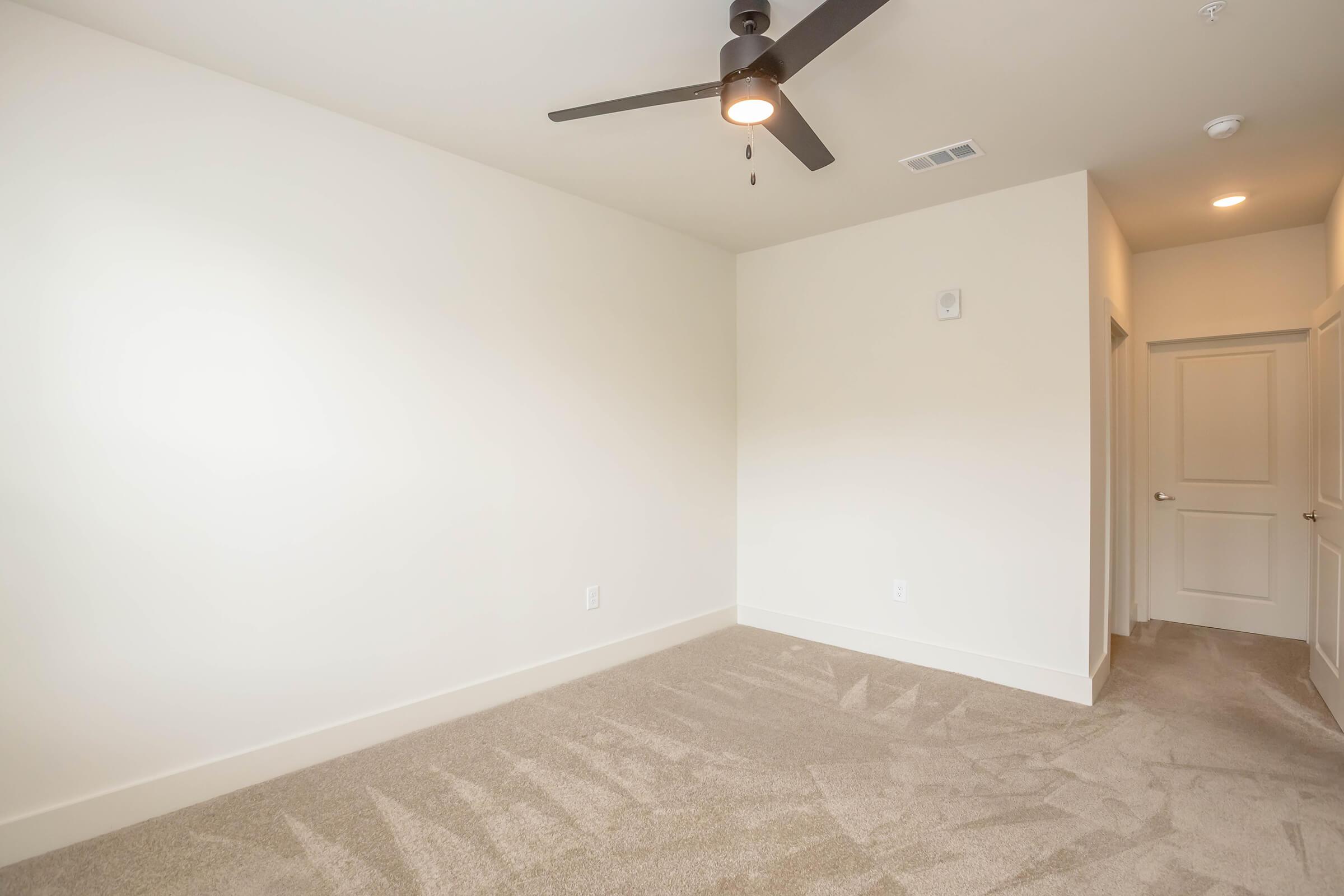 Empty room with light-colored walls and beige carpet. A ceiling fan is installed in the center. One door is visible, leading to another area, while the opposite wall is bare with no furniture or decorations. The space feels bright and uncluttered, perfect for personal customization.