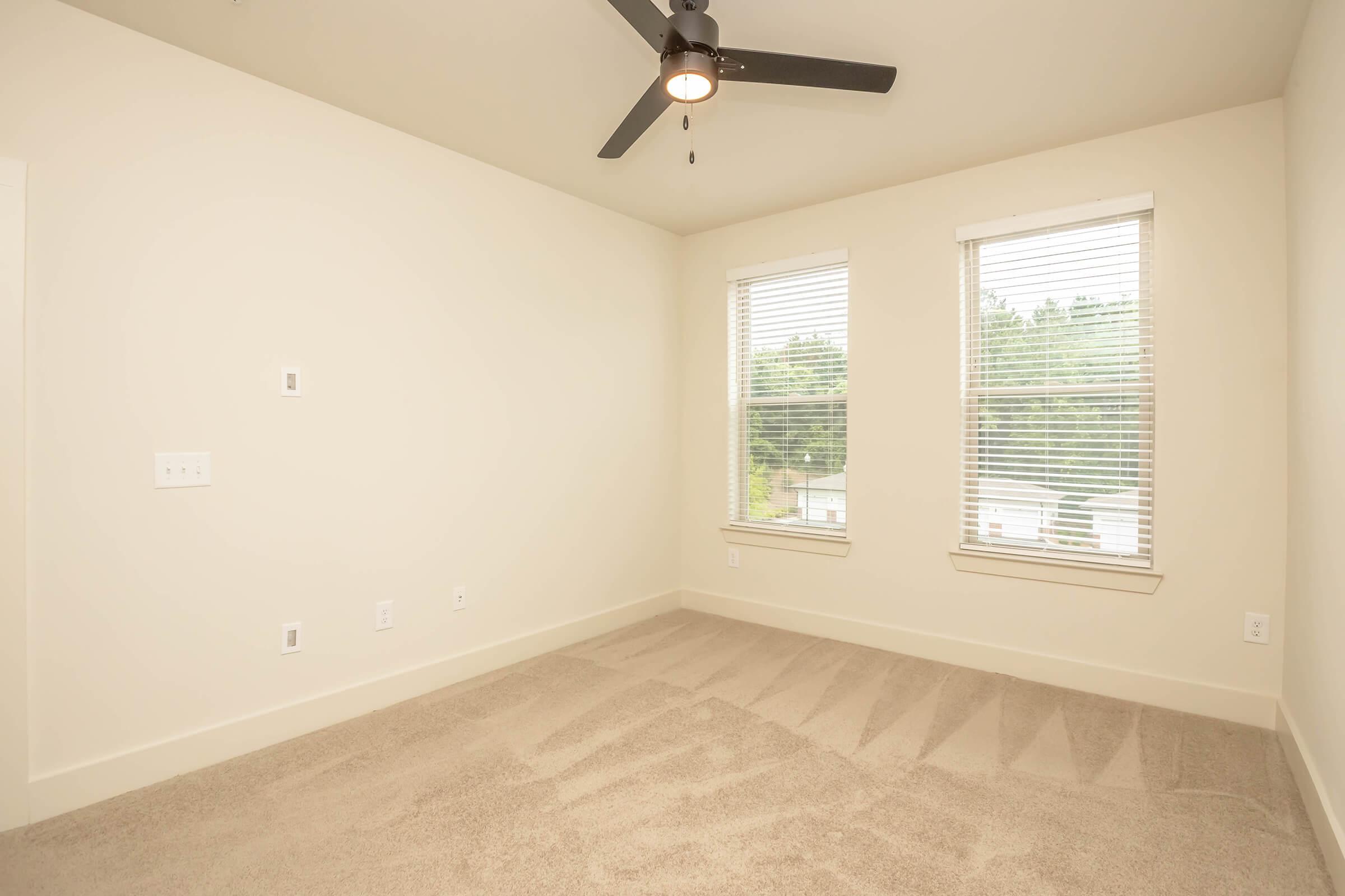 A bright, empty bedroom with light-colored walls and carpet. It features two windows with blinds letting in natural light and a ceiling fan. The room has no furniture, creating a spacious feel.