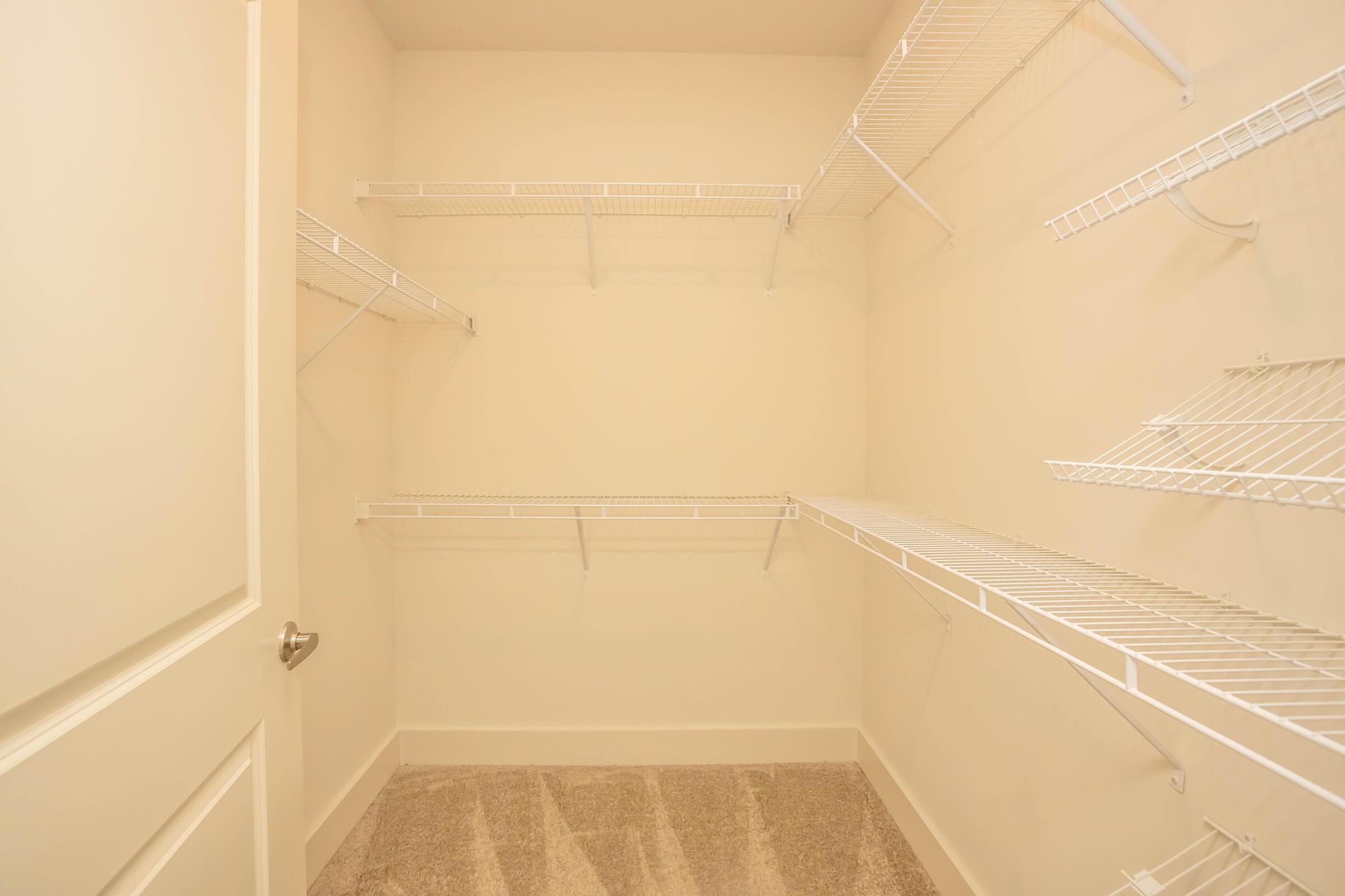 A spacious, empty closet featuring white wire shelving installed on the walls. The closet has a light-colored wall and carpeted flooring, with an open door visible on the left side.
