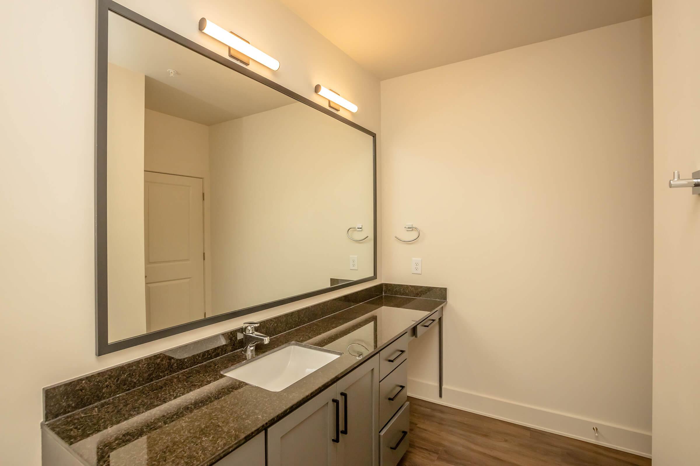 A modern bathroom featuring a spacious dark countertop with a single sink, large wall-mounted mirror, and stylish light fixtures above. The cabinetry is gray, and the walls are a light neutral color. A door is visible in the background, indicating access to another room.