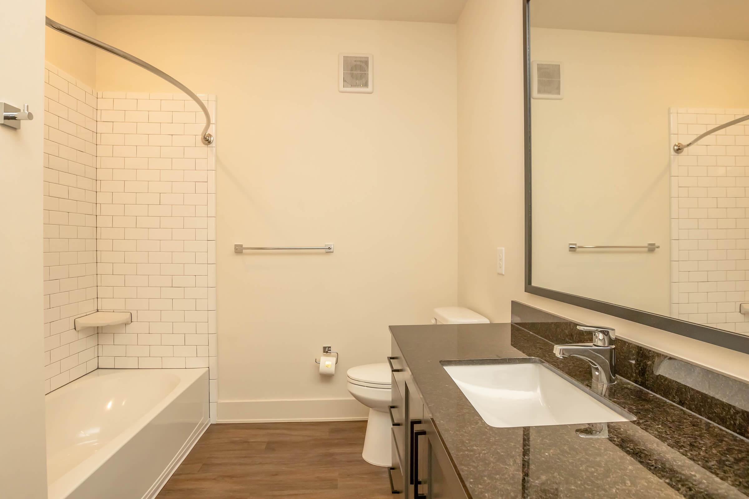 A modern bathroom featuring a bathtub with a white tiled surround, a toilet, and a sleek dark granite countertop with a sink. A large mirror reflects the space, and a towel bar is mounted on the wall. The flooring is a light wood laminate, and the walls are painted a neutral color.