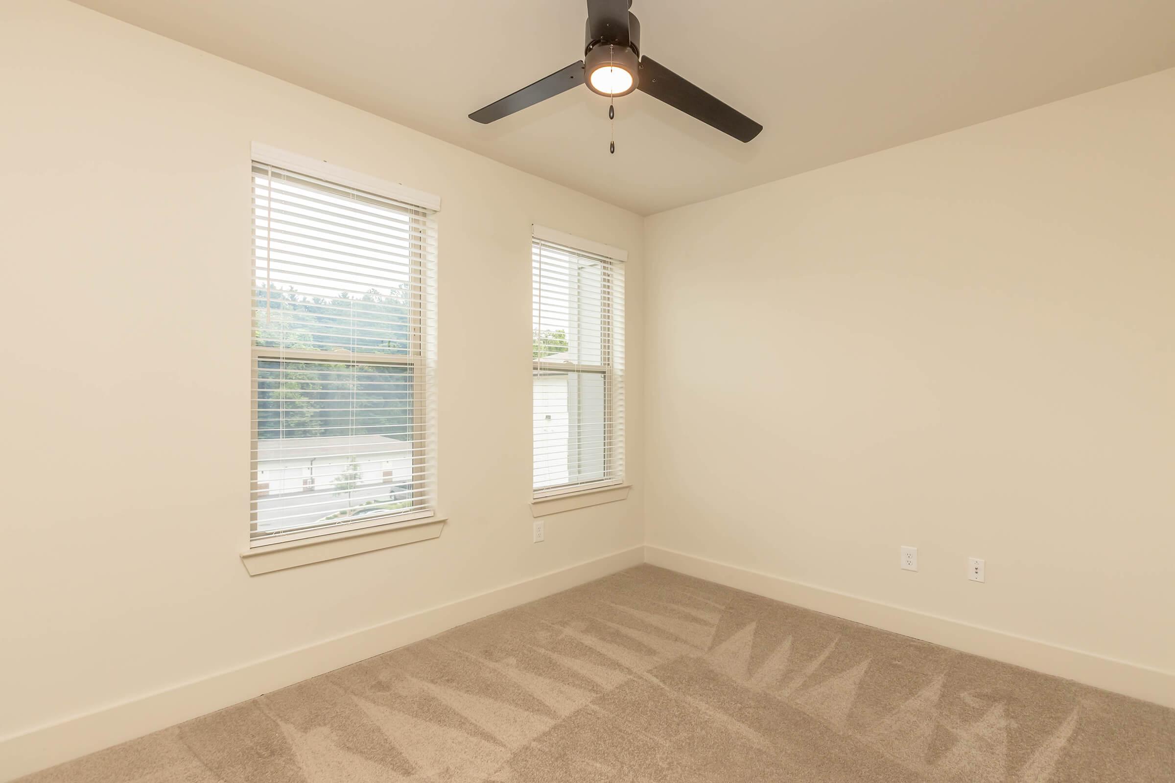 A minimalistic empty bedroom featuring two windows with blinds, a ceiling fan with a light fixture, and light-colored walls. The room has beige carpet flooring and an overall bright and airy atmosphere.