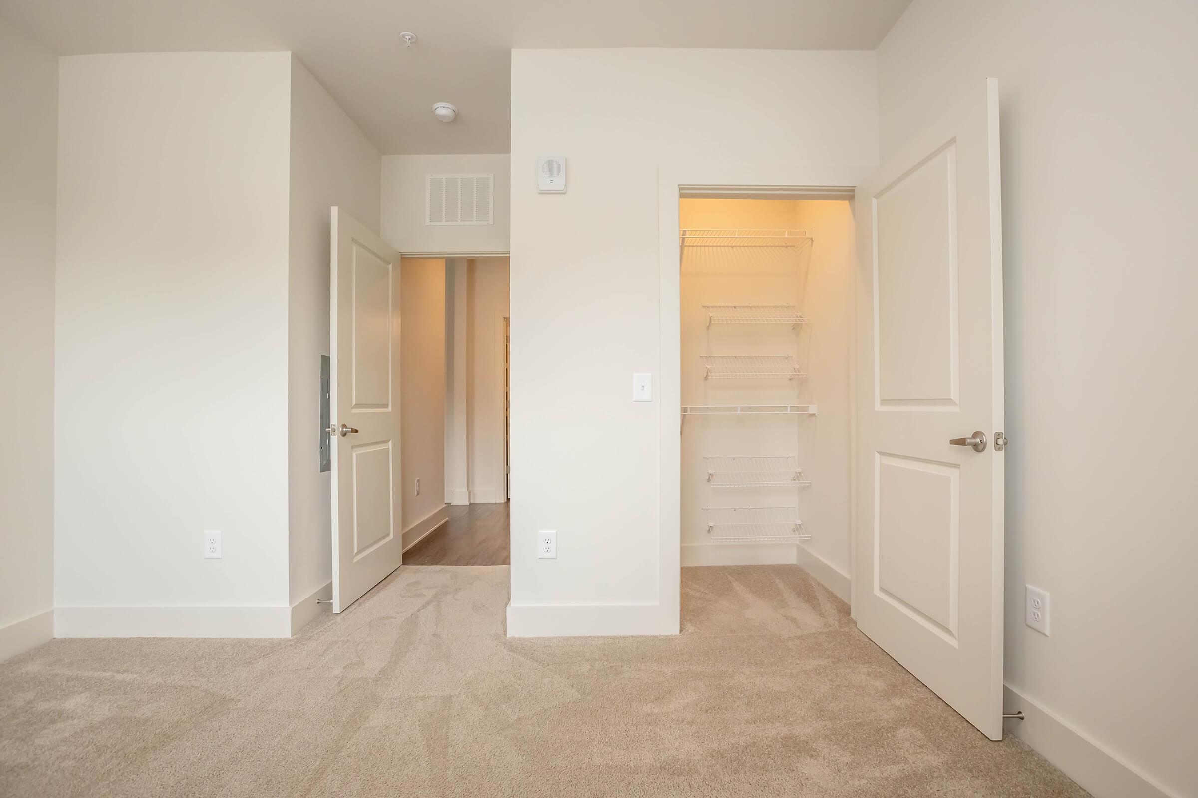 A light-colored room with beige carpet, featuring two open doors. One door leads to a closet with wire shelving, while the other door opens into another room. The walls are painted in a soft hue, enhancing the spacious and airy feel of the area.